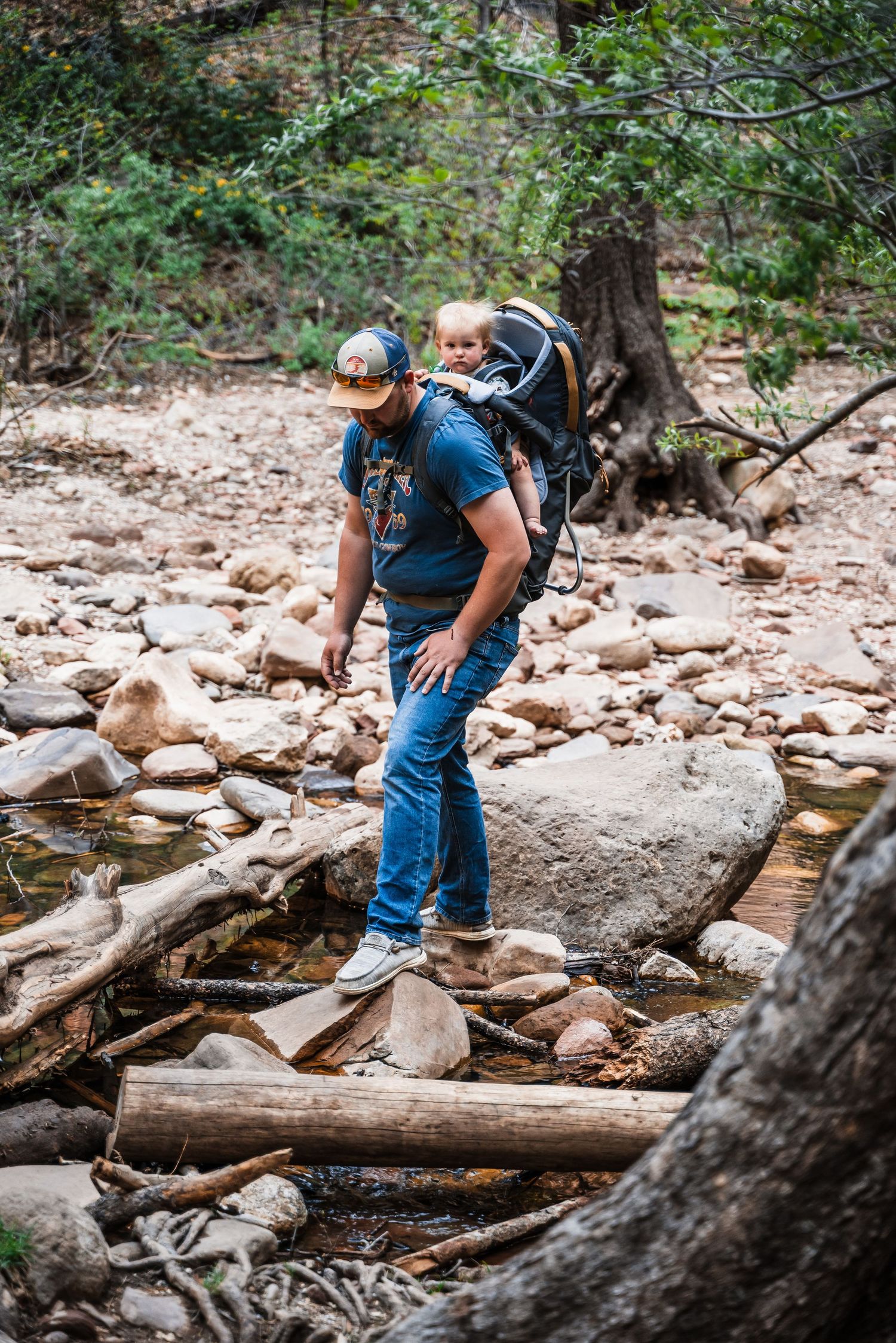Hiker crossing a rocky stream bed with hiking gear and backpack in a wooded area.