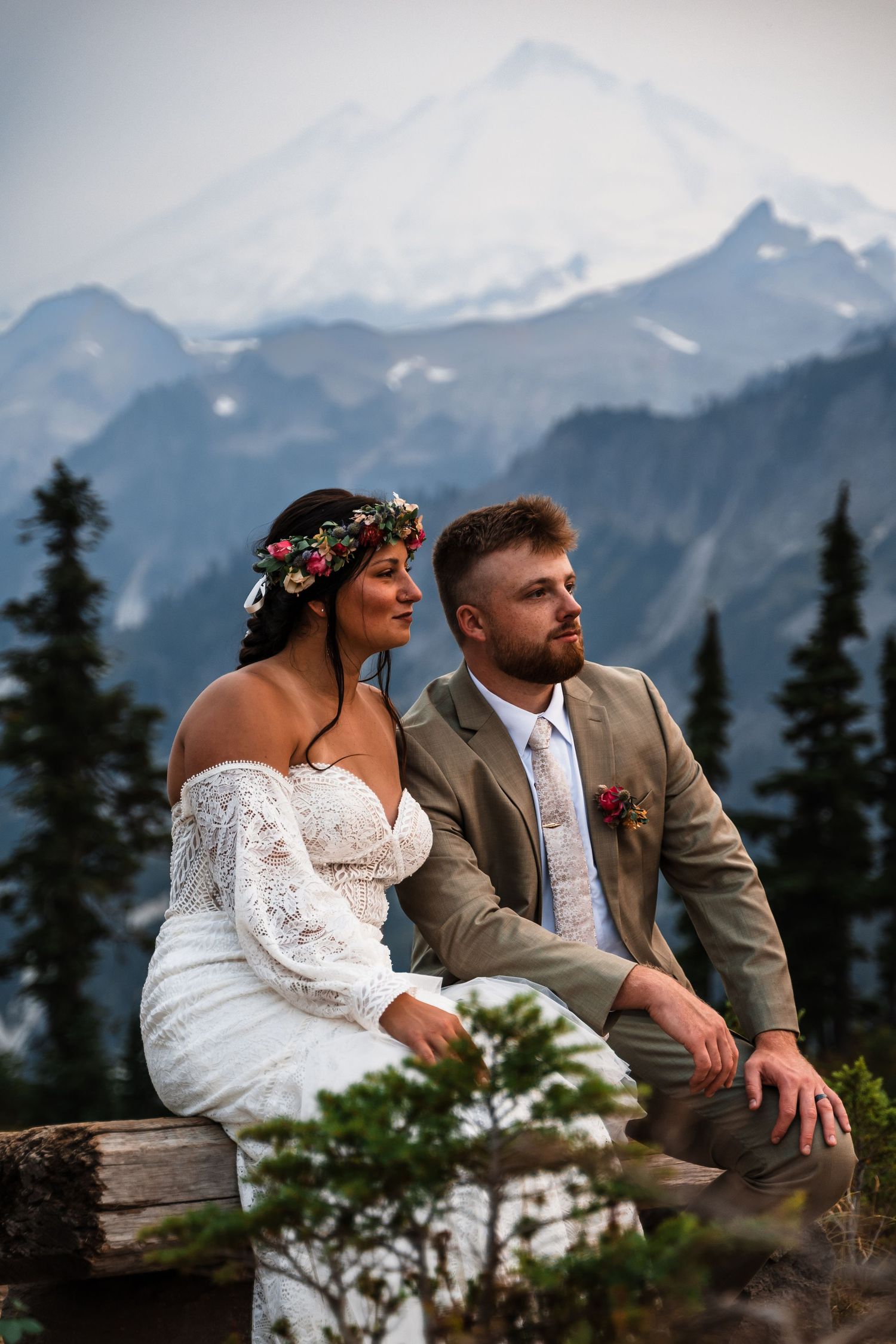 A couple sits on a rustic bench overlooking snow-capped mountain peaks in a romantic mountain setting.