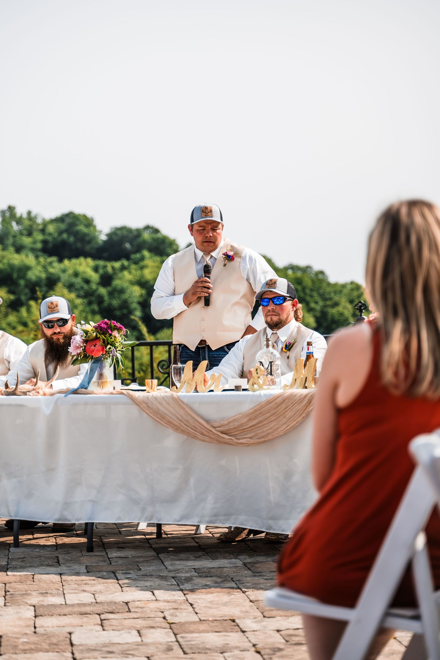 A wedding toast being given at an outdoor reception with guests seated at a decorated head table on a stone patio.