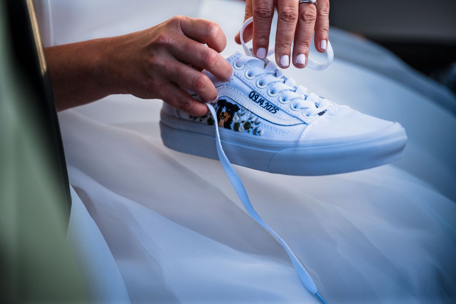 Close-up of white wedding sneakers being laced up against flowing bridal dress fabric.