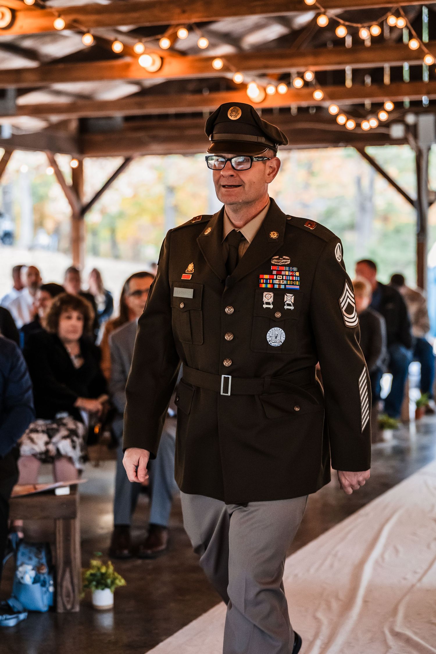 Military service members in dress uniforms gather at an indoor event space with string lights overhead.