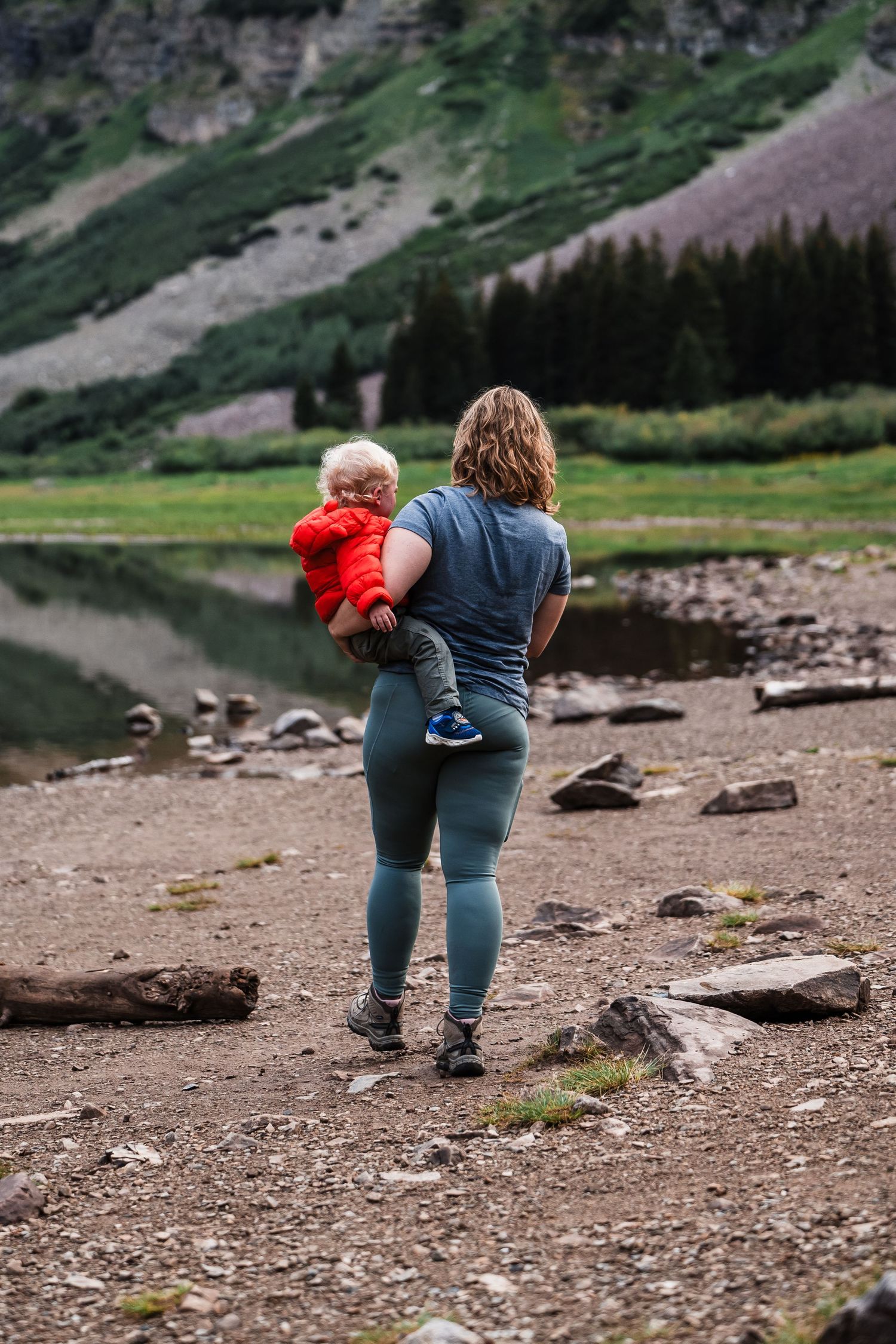 Adult and child standing by mountain lake shore with dramatic peaks in background.