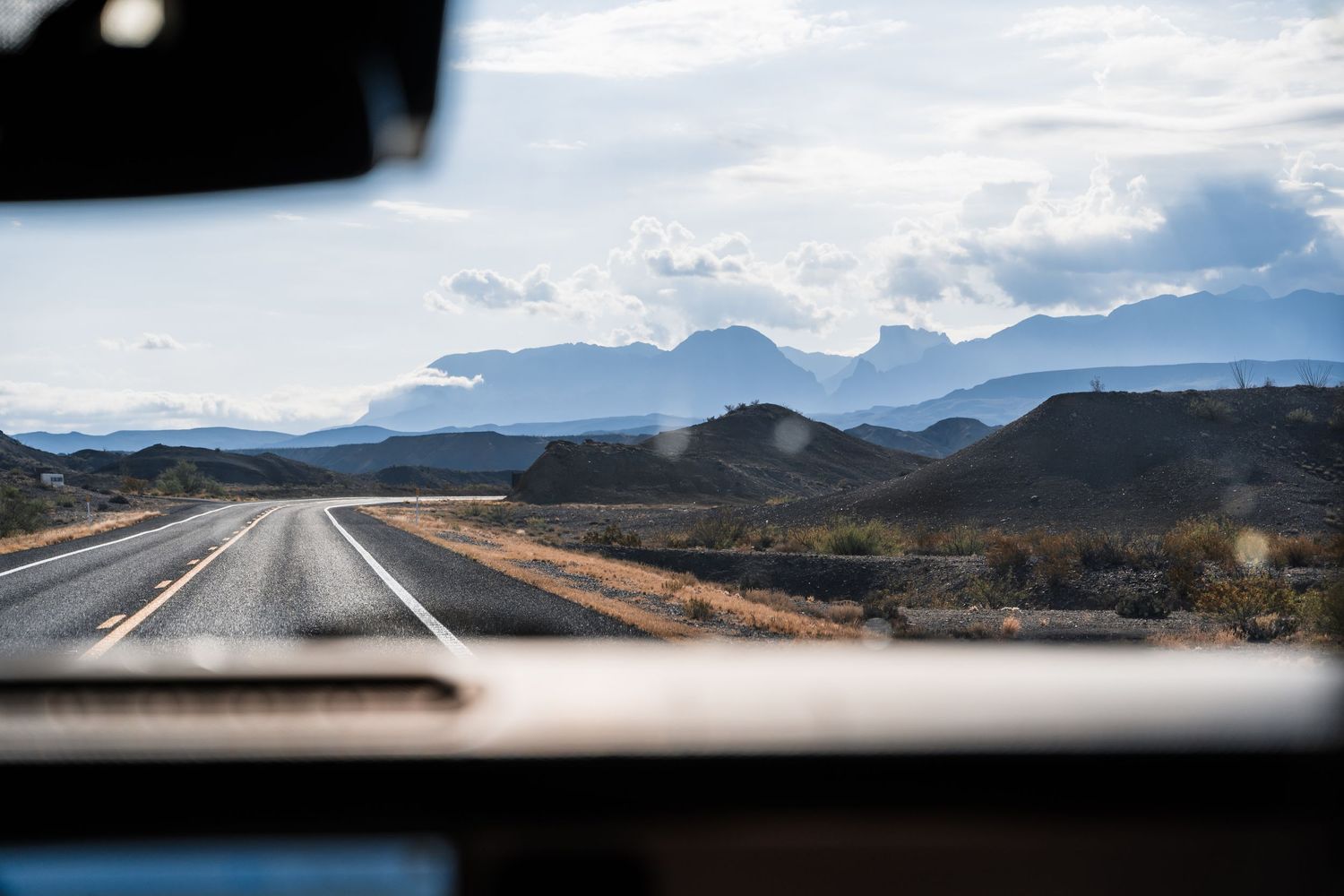 A scenic highway stretches towards distant mountains under cloudy skies, viewed through a car windshield.