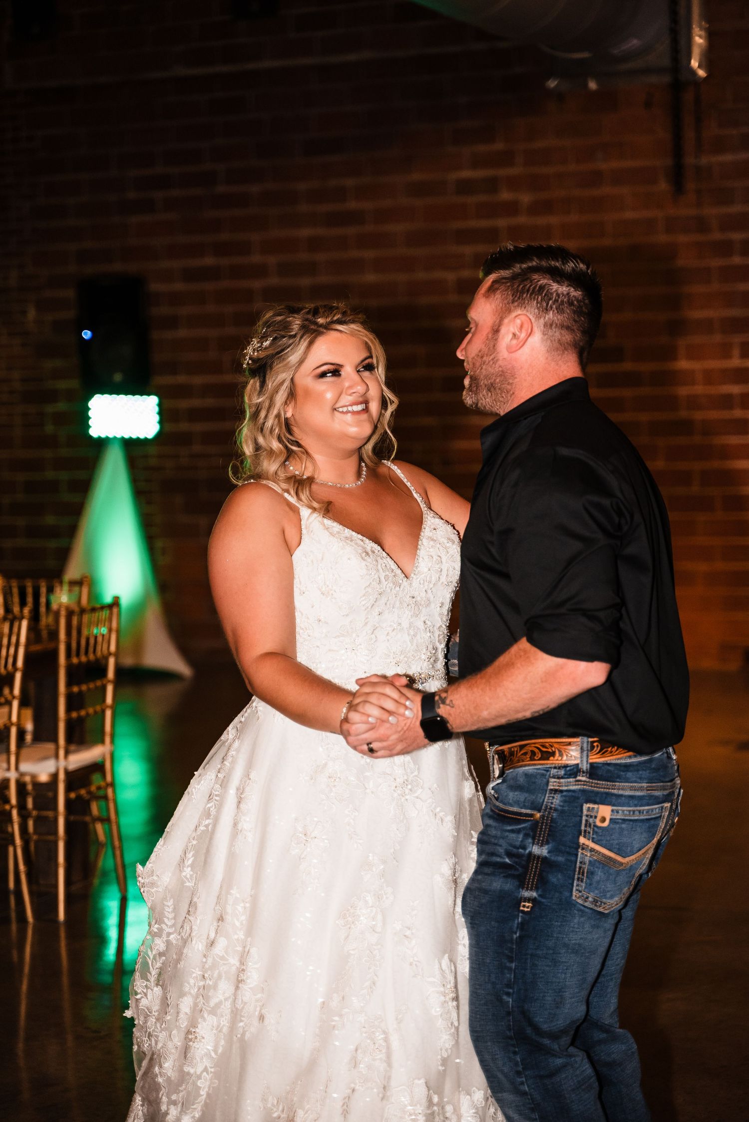 A couple dances together at a wedding reception with exposed brick walls and green uplighting in the background.