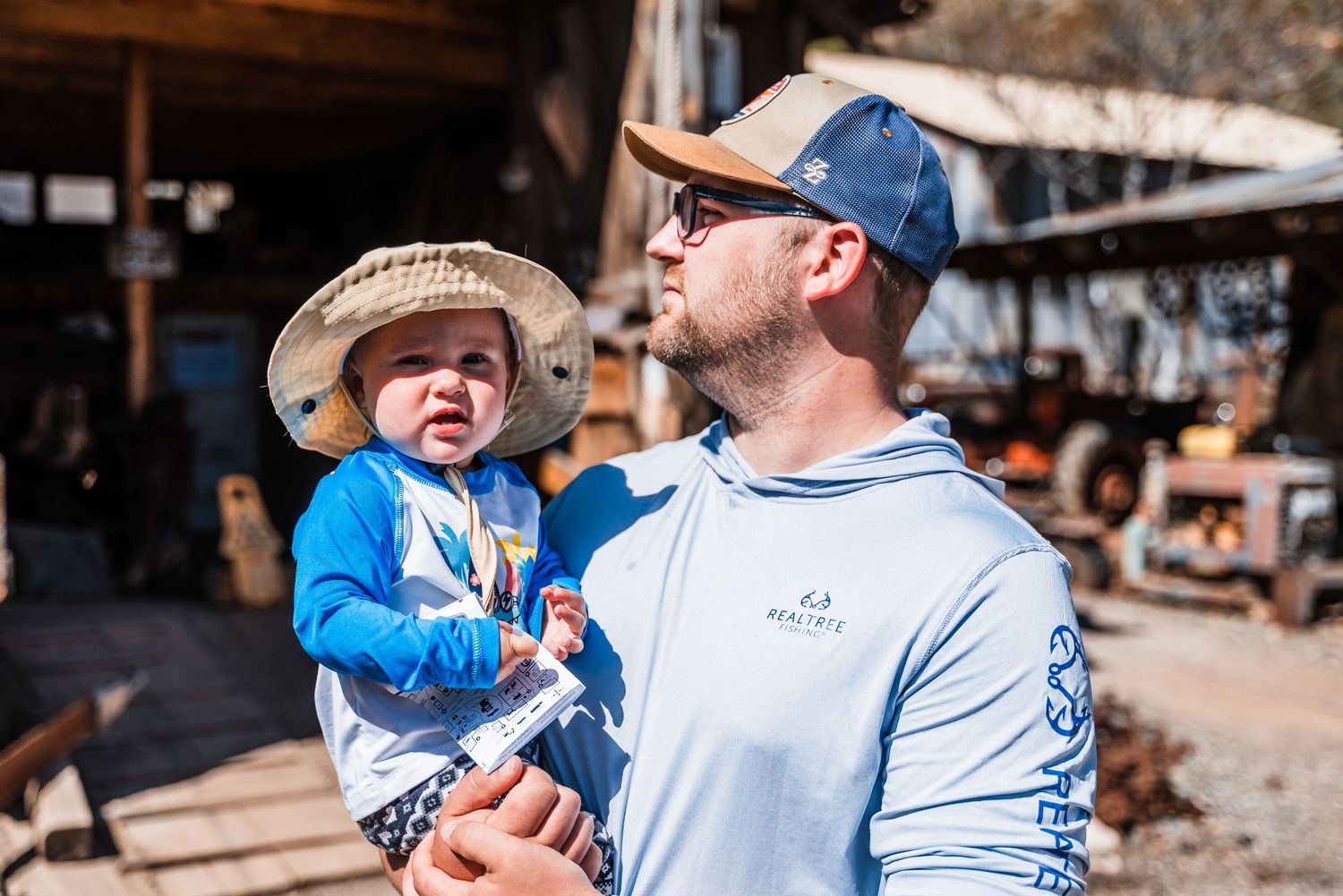 A young child wearing blue and an adult in a baseball cap stand together outdoors by a rustic wooden structure.