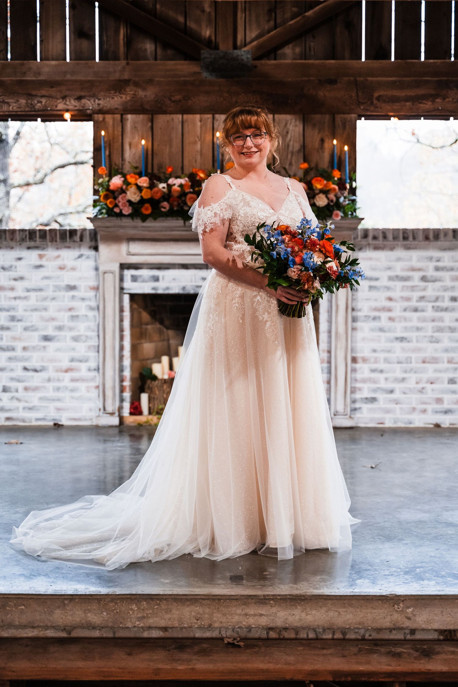 Bride holding vibrant blue and orange bouquet poses in front of rustic fireplace.