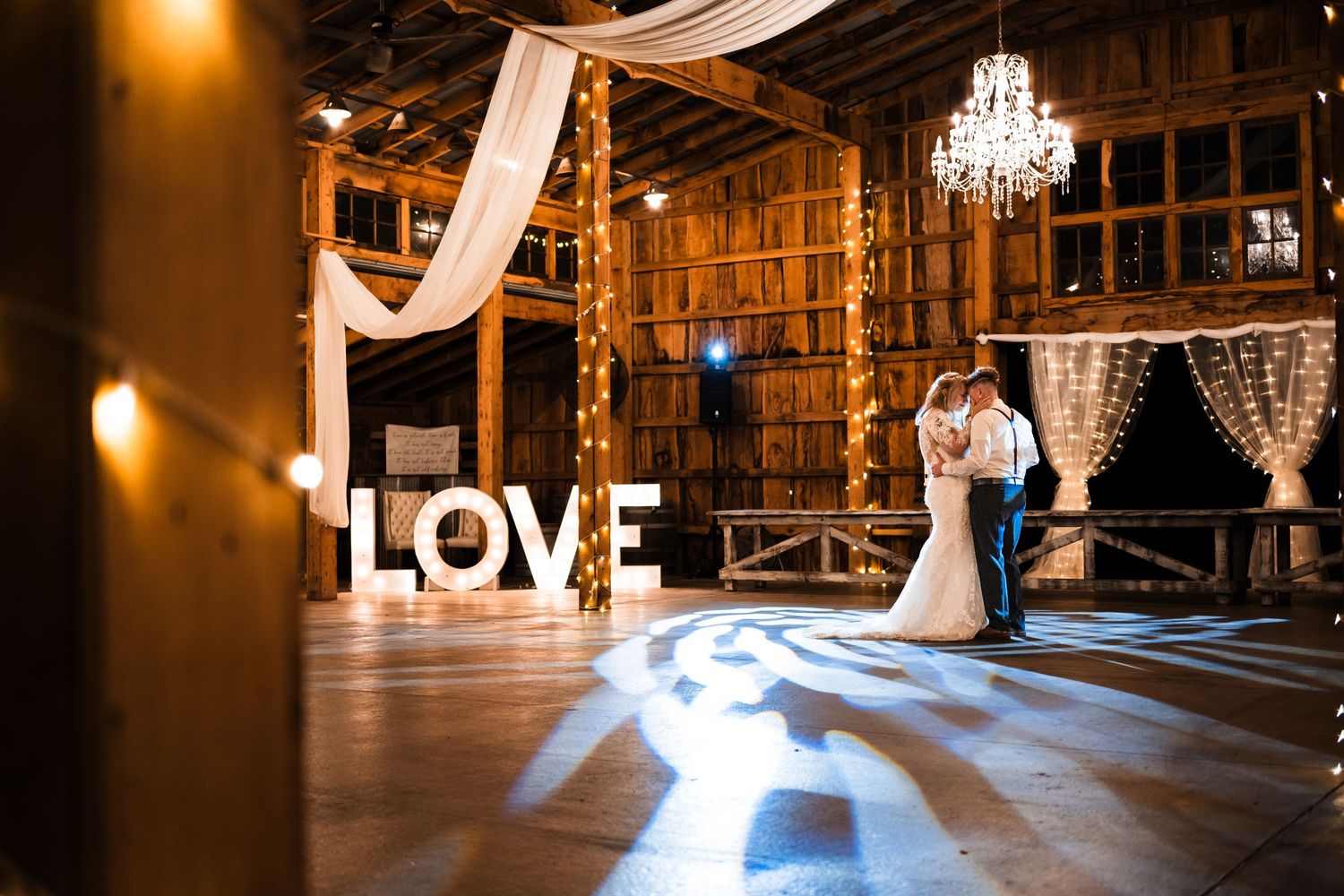 Rustic barn wedding reception with illuminated LOVE sign, chandelier, and couple dancing on wooden floor in warm lighting.