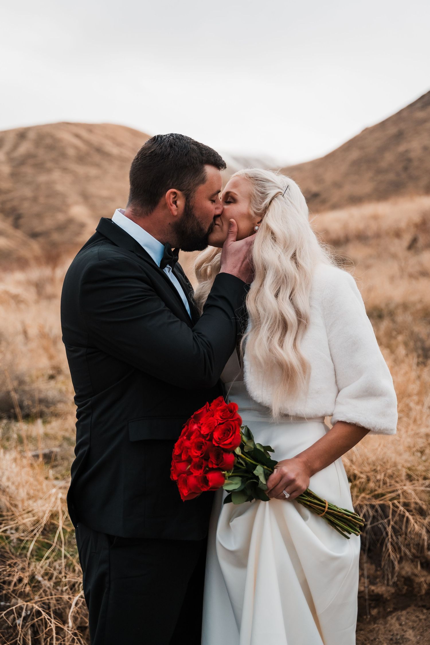 Intimate moment between newlyweds with red roses against desert mountain backdrop.