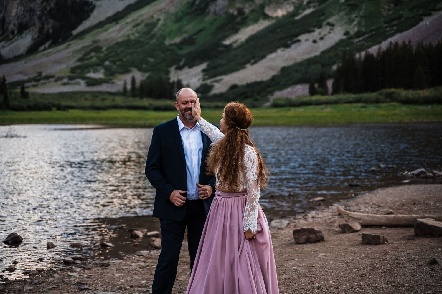 A couple stands by a mountain lake surrounded by majestic rocky peaks and alpine scenery.