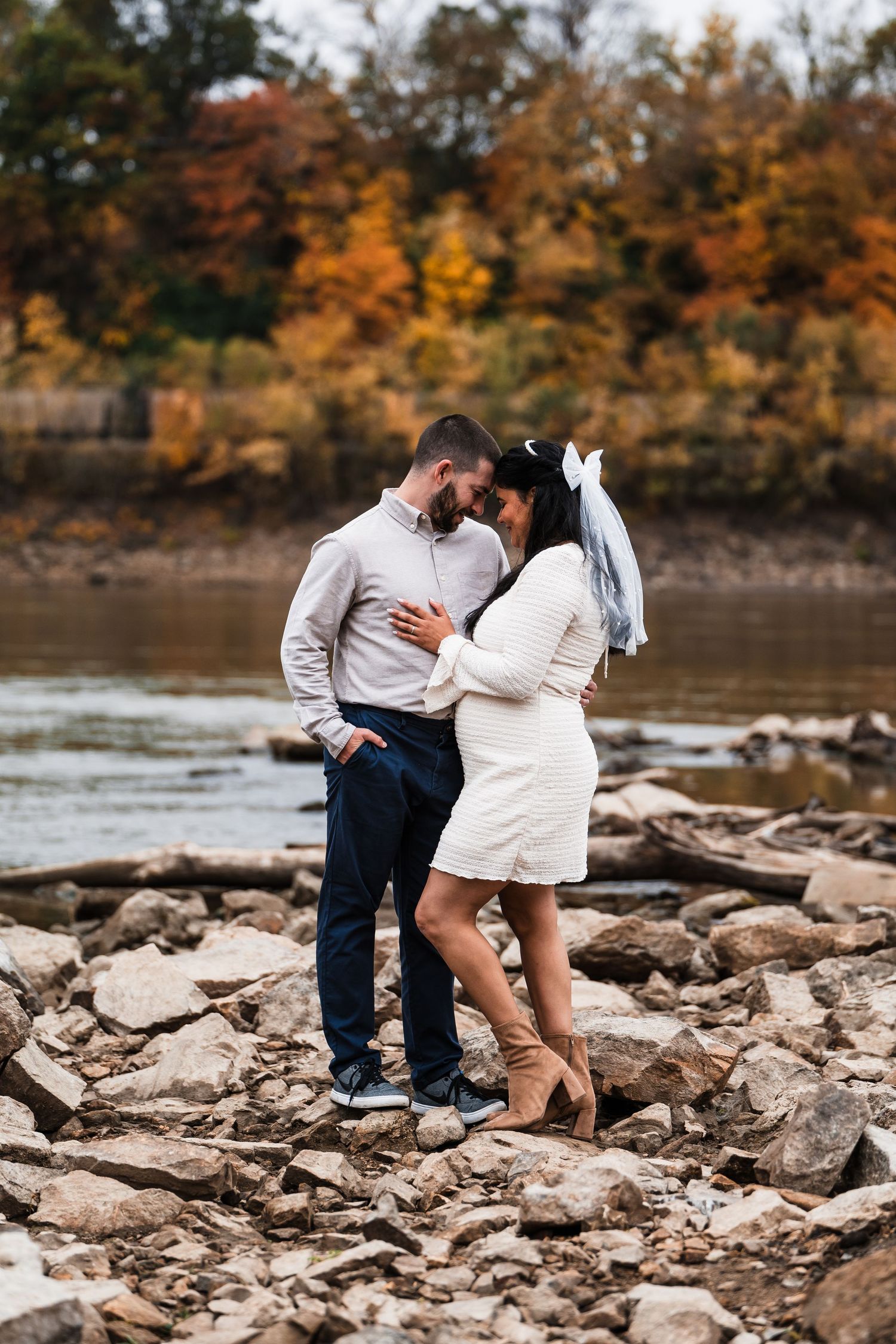 A couple sharing an intimate moment on rocky riverbank with fall foliage in background.