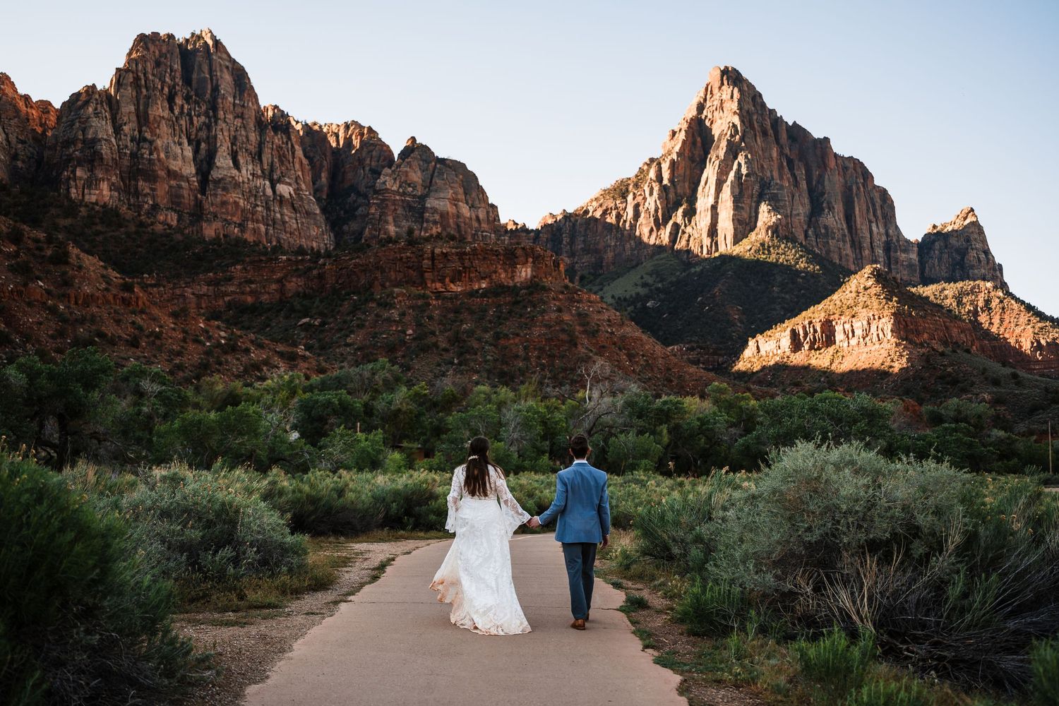 Bride and groom walking hand in hand toward majestic Zion National Park canyon at dusk.