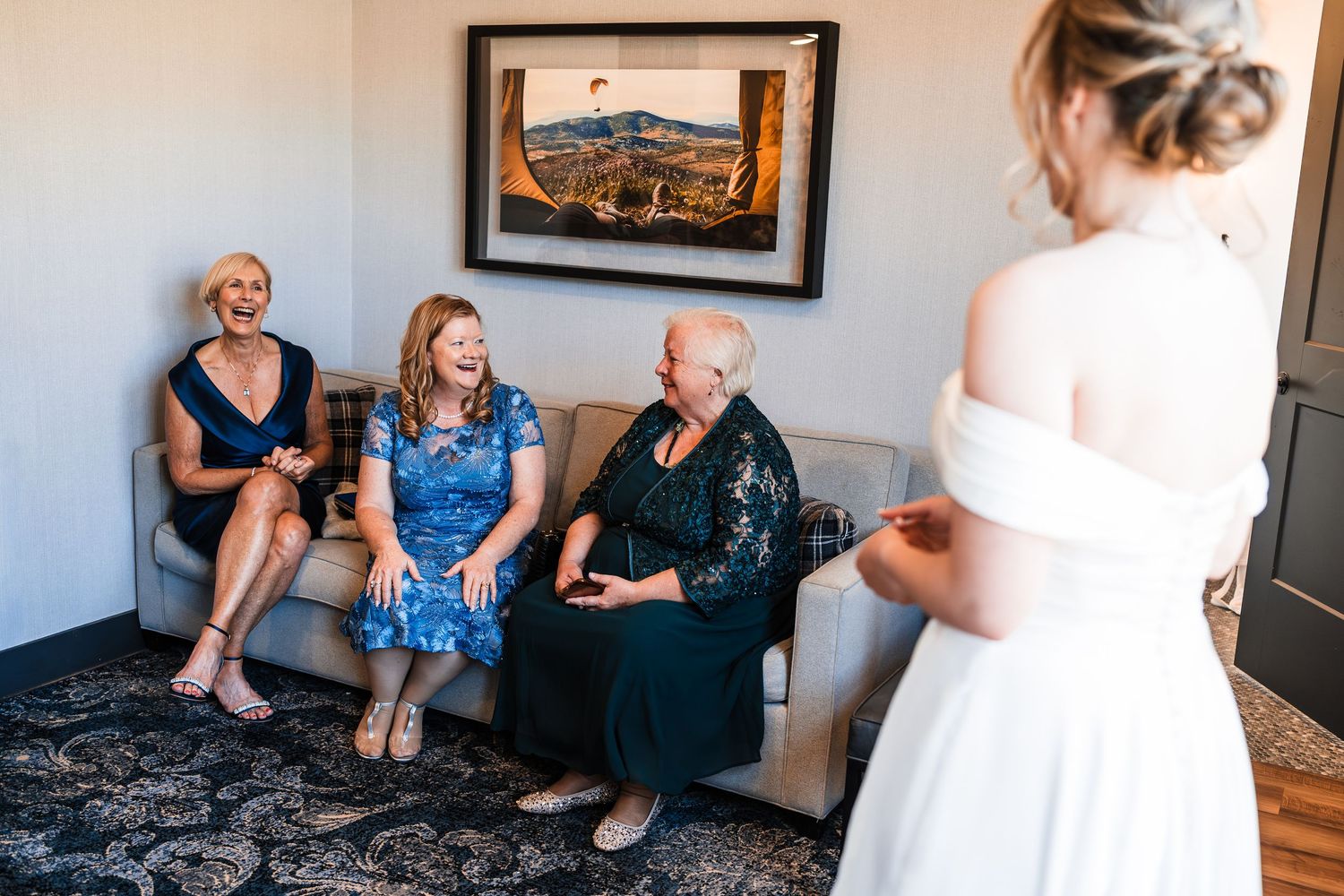 Bride in white gown reveals dress to smiling family members seated on a couch in a warmly decorated room.