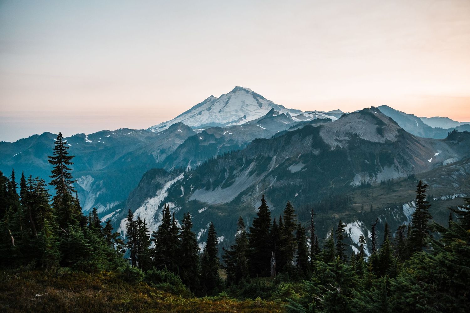 A panoramic mountain landscape with snow-capped peaks and evergreen forests during sunset in the Pacific Northwest.