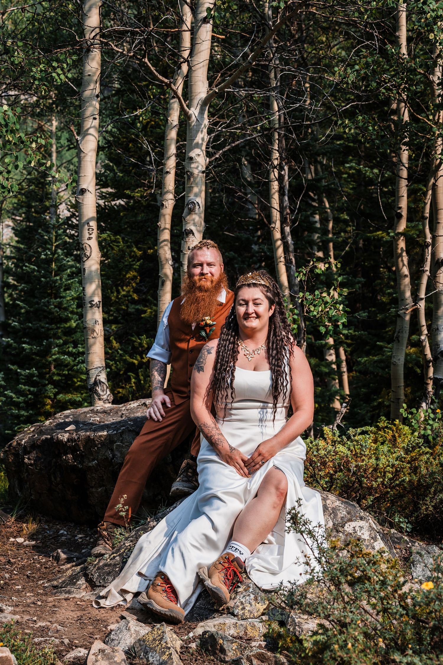 Tattooed bride and groom pose on forest rocks surrounded by aspen trees in a woodland wedding portrait.
