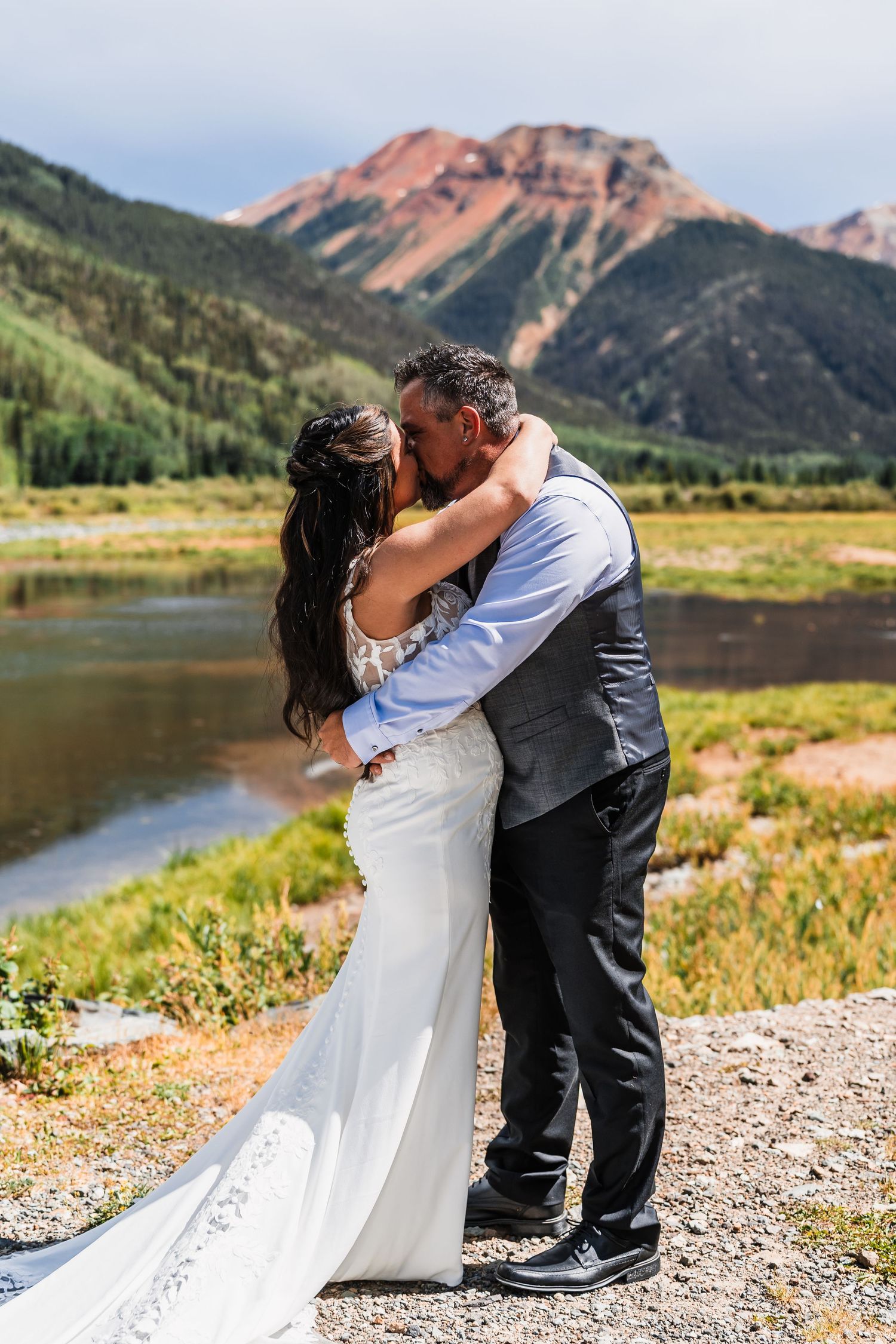 Bride and groom kiss romantically with a stunning red mountain and river landscape behind them.