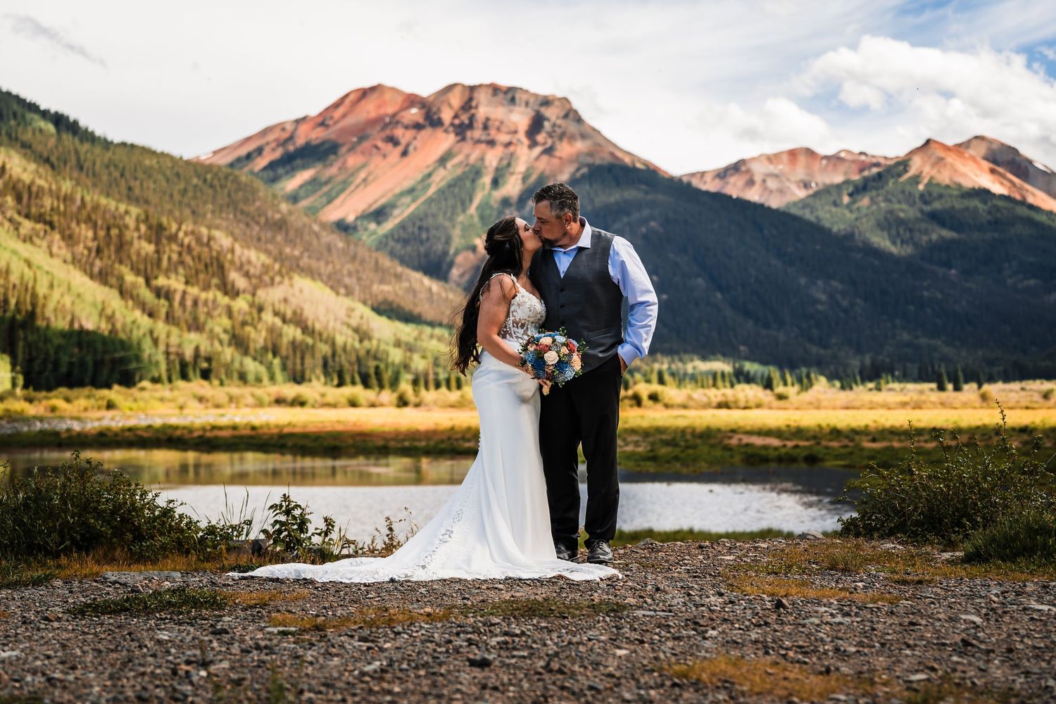 Bride and groom share a romantic kiss in front of a stunning mountain lake backdrop during their outdoor Colorado wedding.