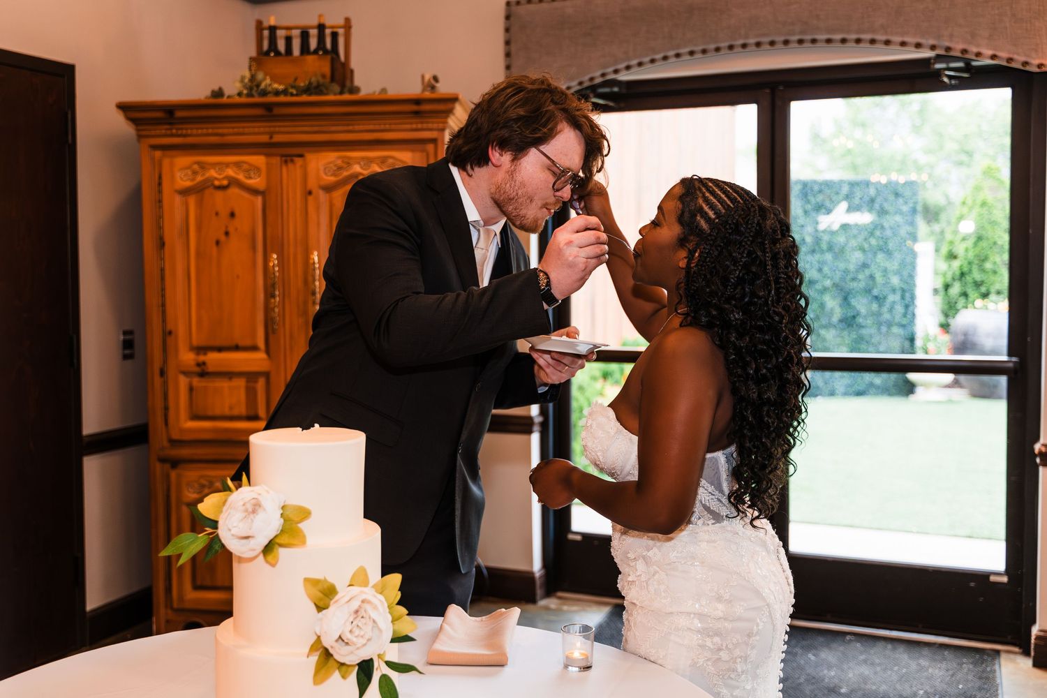 Bride and groom sharing wedding cake at their reception, smiling intimately near a white tiered cake.