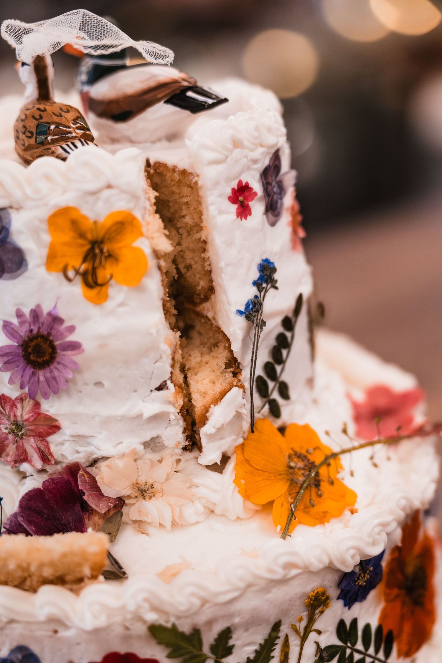 Three-tier white wedding cake decorated with colorful pressed flowers and bird topper.
