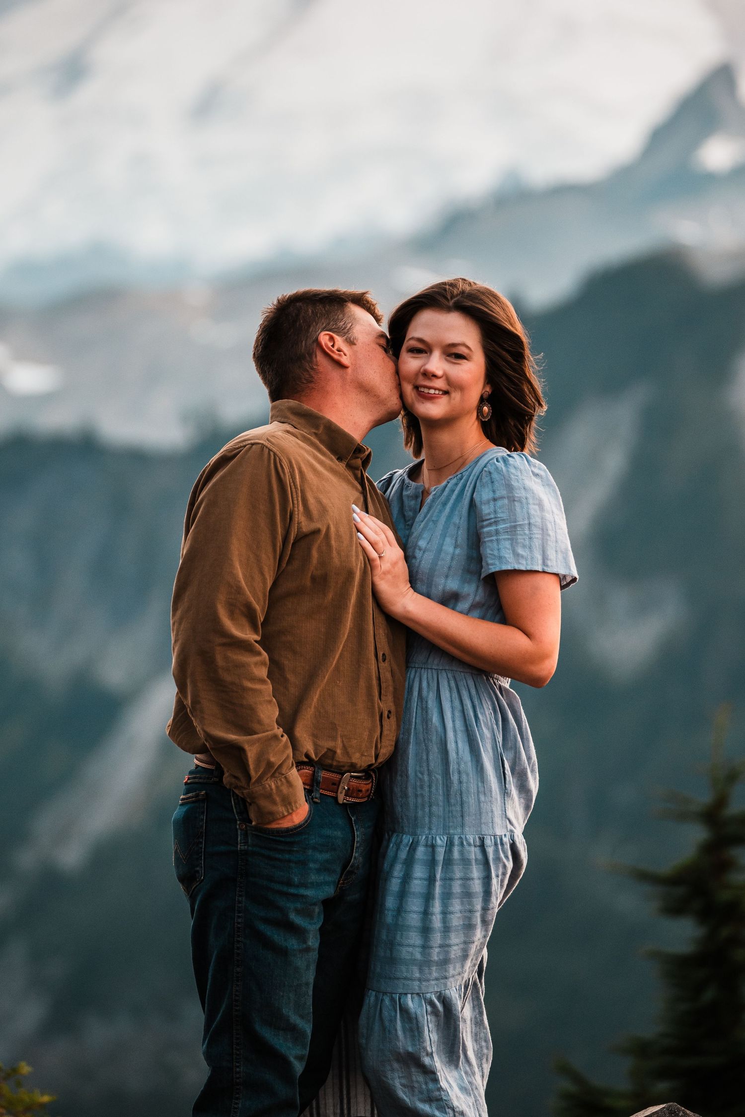 A romantic couple embraces against a backdrop of majestic mountain peaks on a cloudy day.