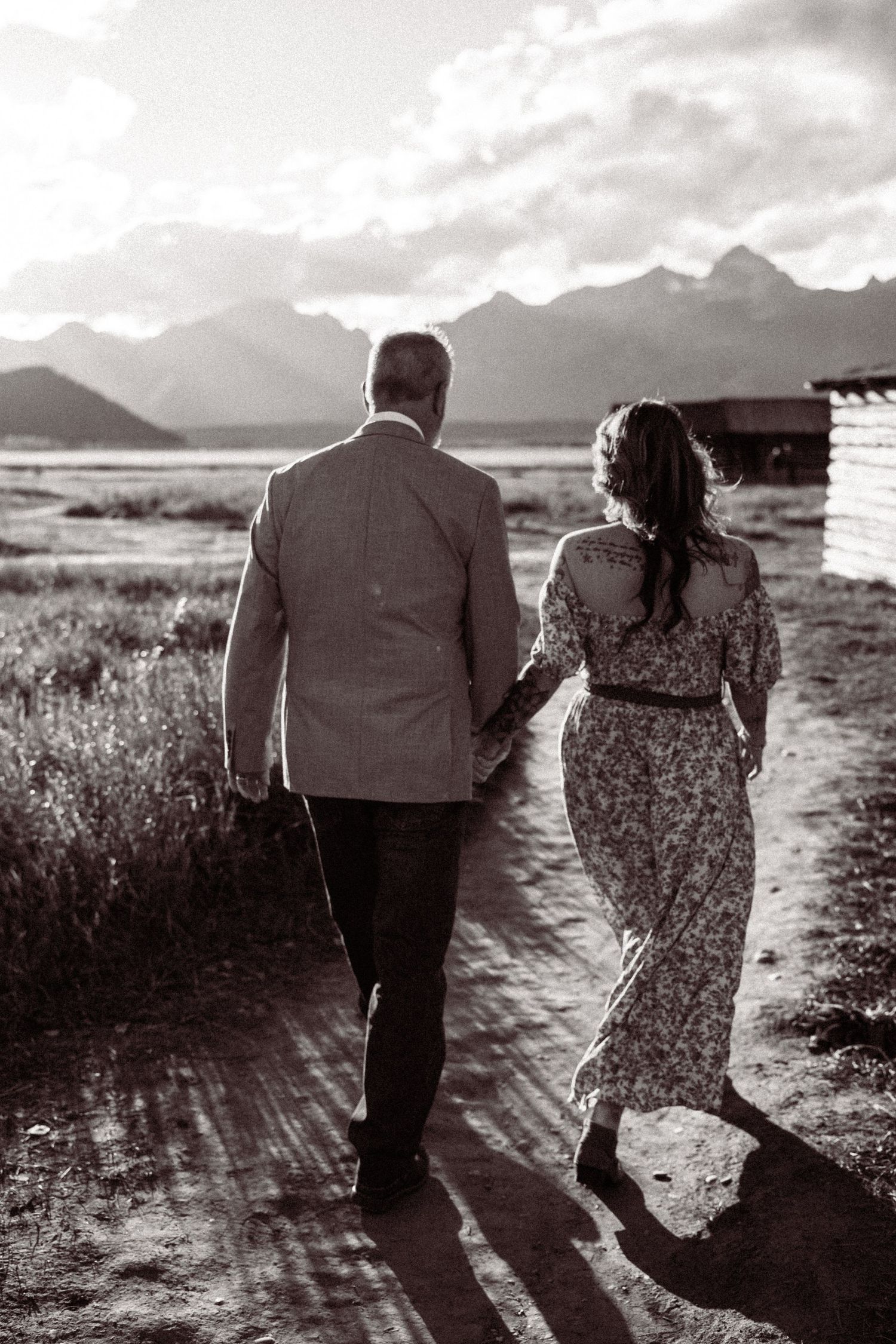 A couple walks hand in hand along a scenic mountain path in black and white photography.