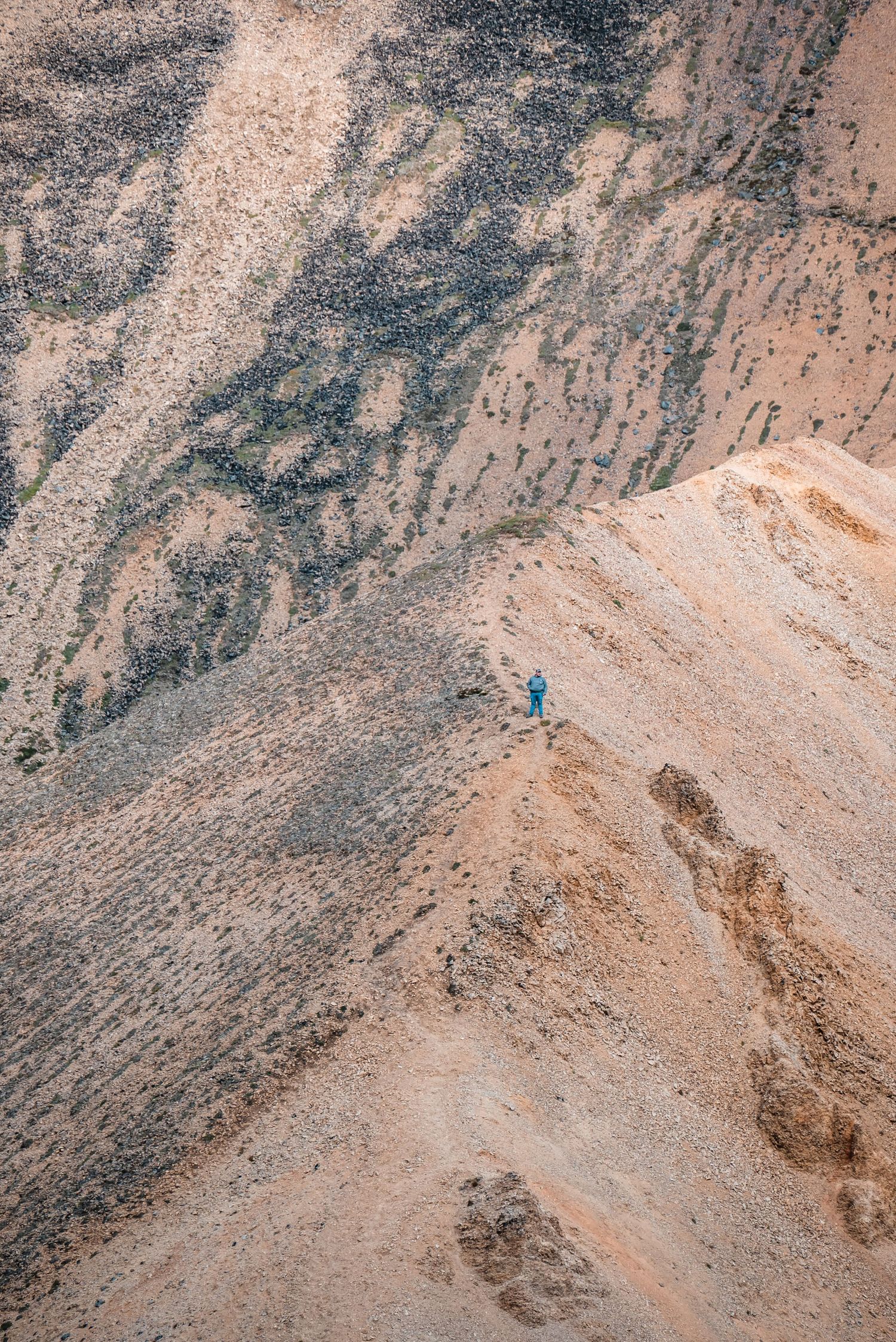 A tiny hiker traverses a steep mountainous trail on a rugged desert landscape.
