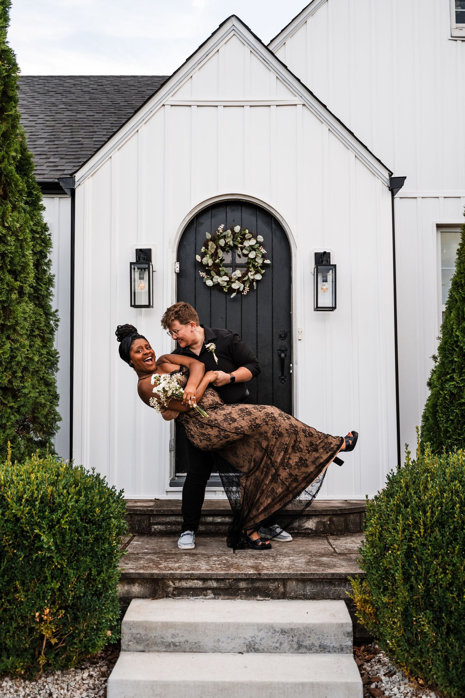A couple posing playfully on the steps of a white chapel with a black door and wreath.