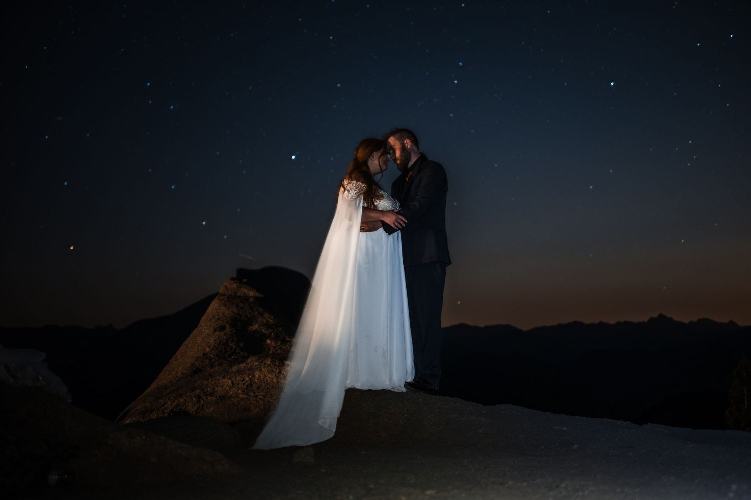A romantic silhouette against a starry night sky stands atop a mountain peak at twilight.