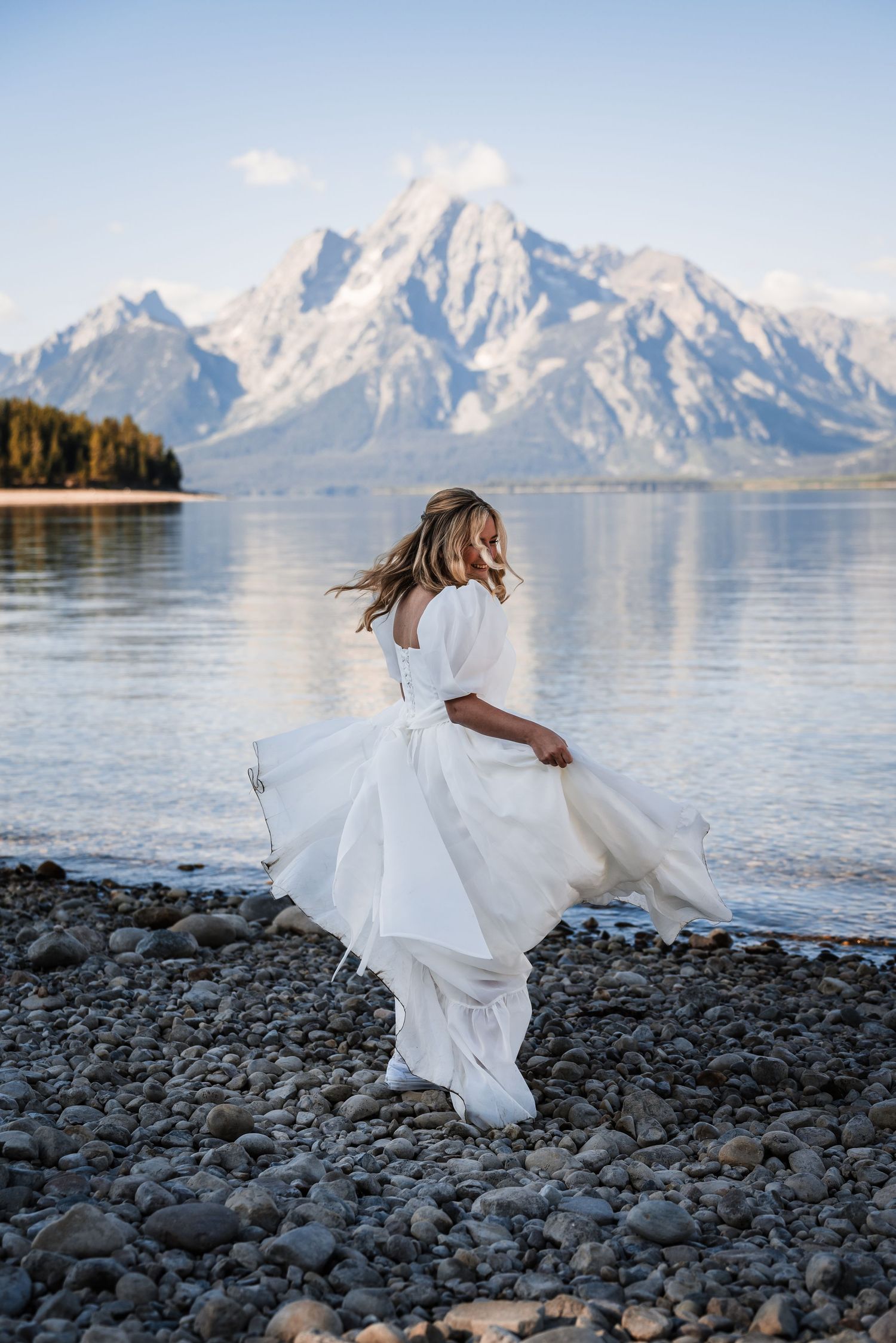 A flowing white wedding dress twirls against a dramatic mountain lake backdrop.