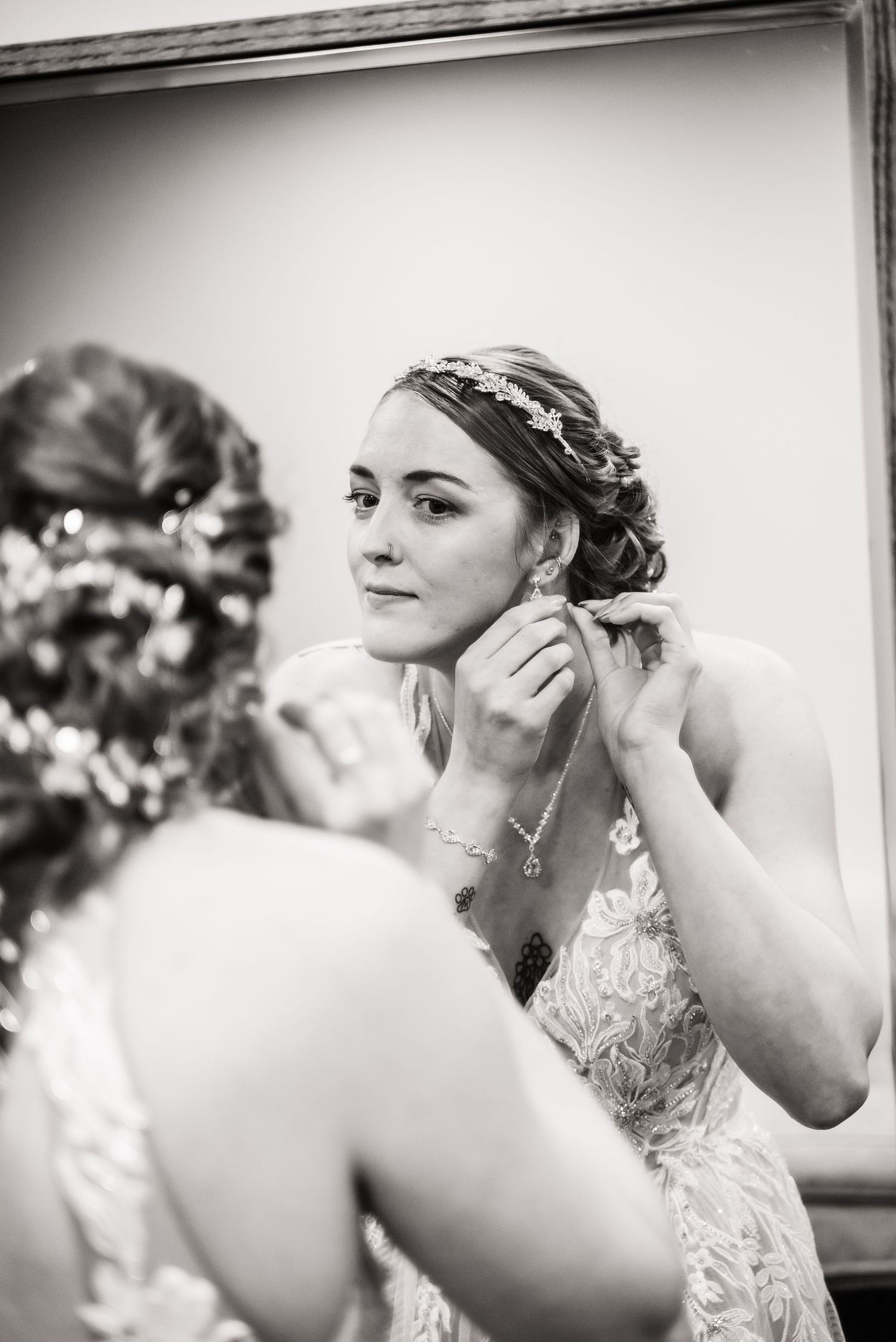 A bride putting on earrings while looking in a mirror wearing a lace wedding dress with an updo hairstyle.