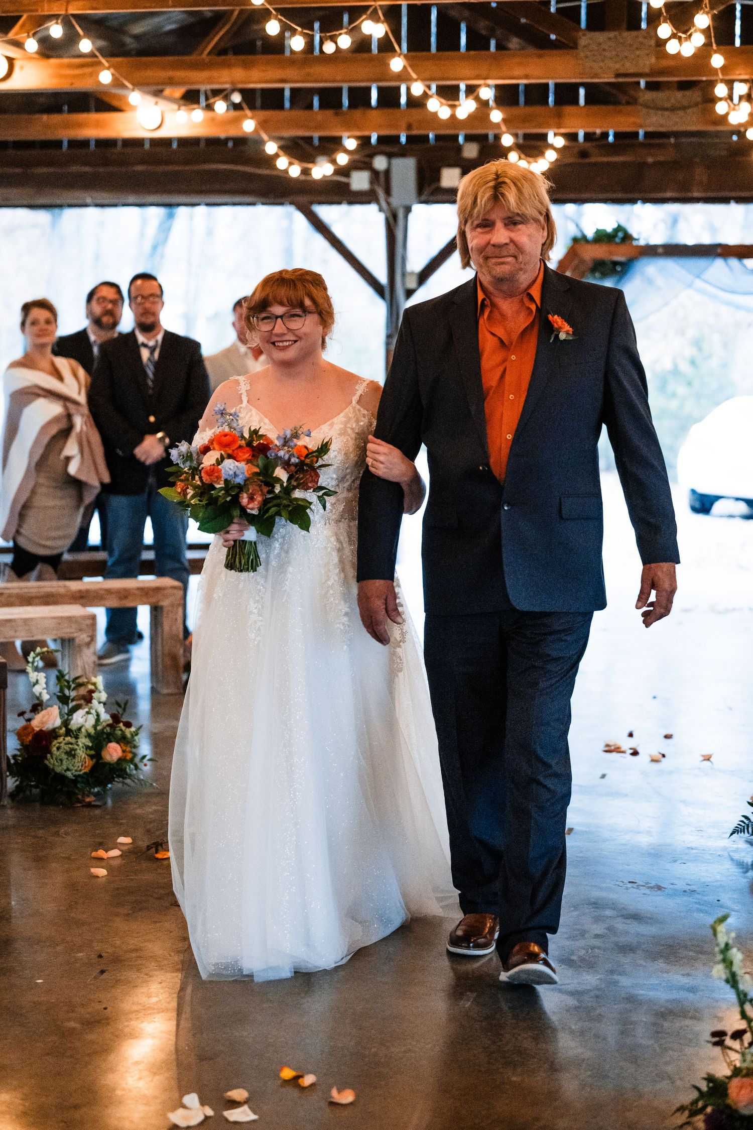Wedding ceremony in a rustic venue decorated with string lights as two people walk down the aisle.