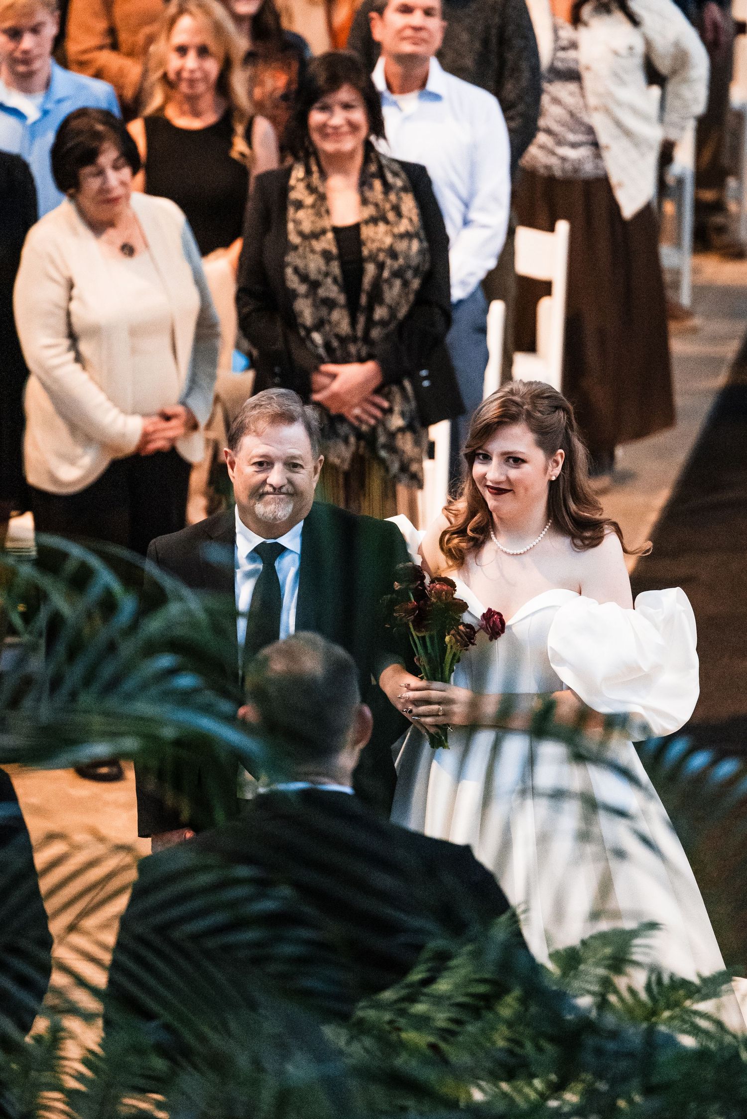 Wedding ceremony taking place in a tropical indoor setting with palm fronds and guests watching.