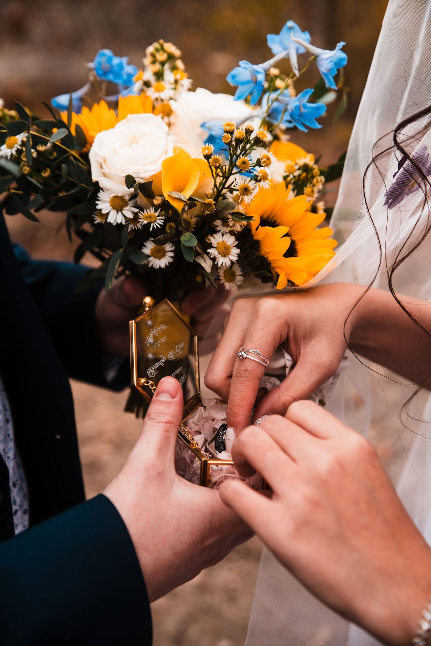 Hands hold a wedding bouquet featuring sunflowers and blue delphinium flowers.