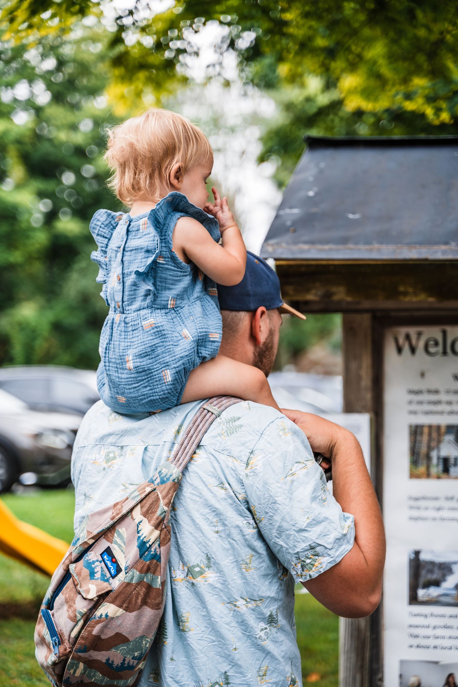 A child wearing denim sits on someone's shoulders while looking at an outdoor information board.