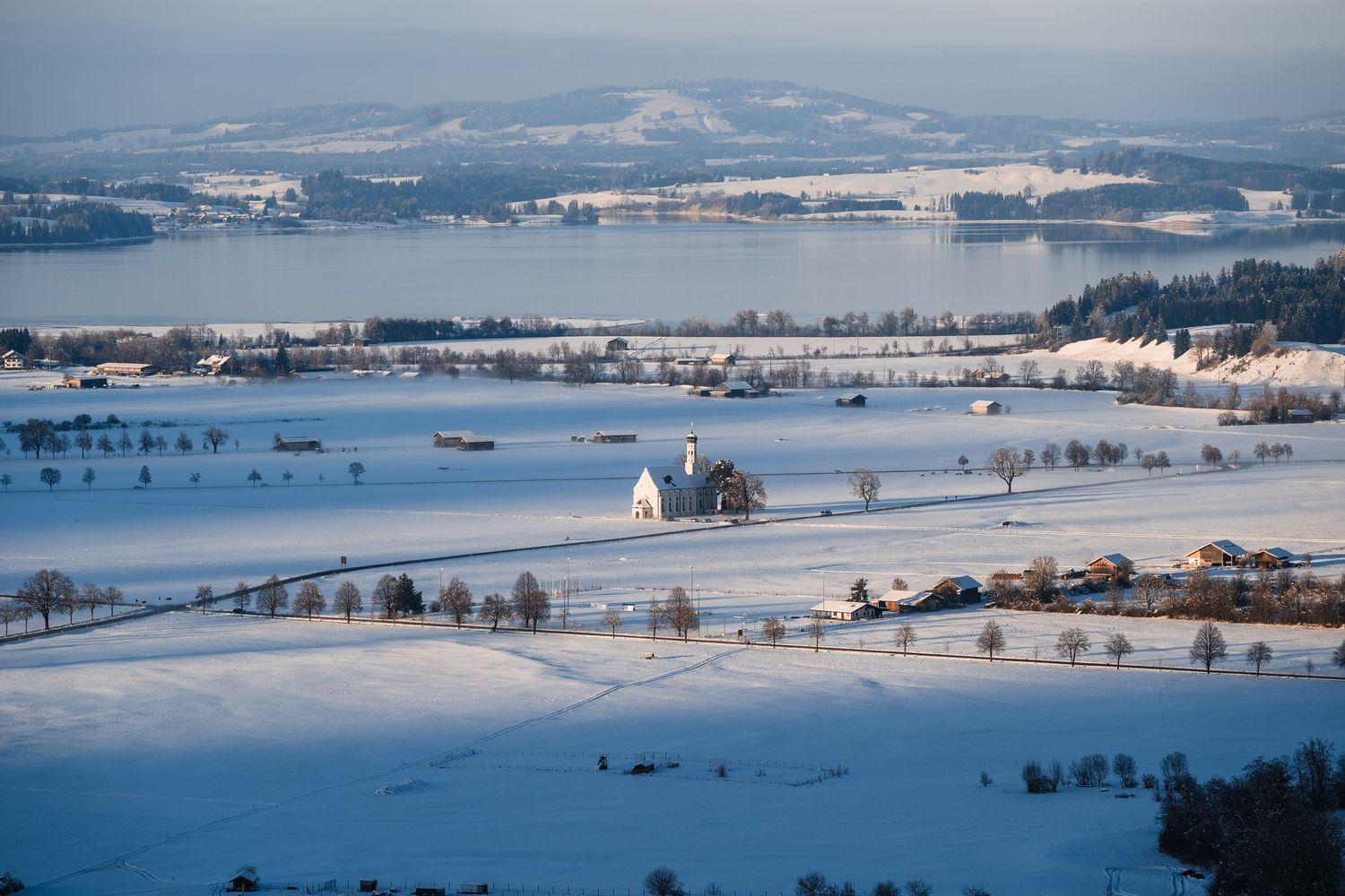 Panoramic winter landscape of snow-covered fields and distant mountains under a pale blue sky.