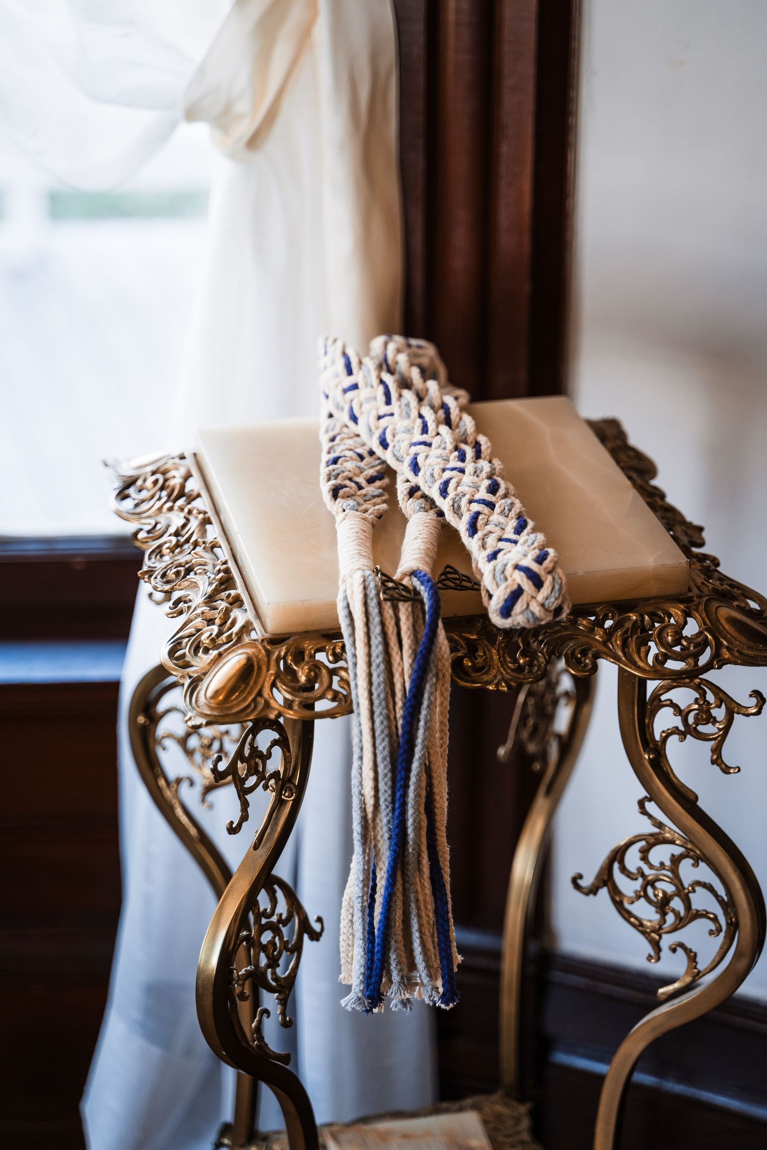 Decorative braided rope handfasting cord with blue and cream tassels resting on an ornate gold table.