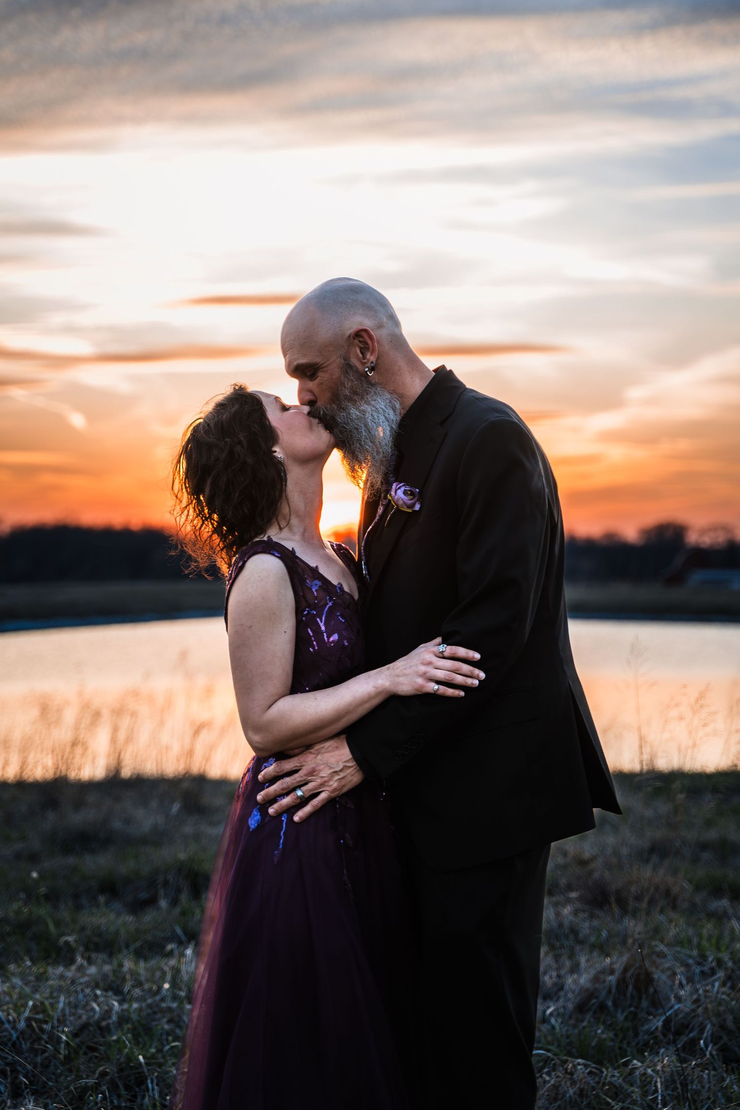 Couple sharing romantic sunset kiss near lake, bride in purple gown, groom in black suit.