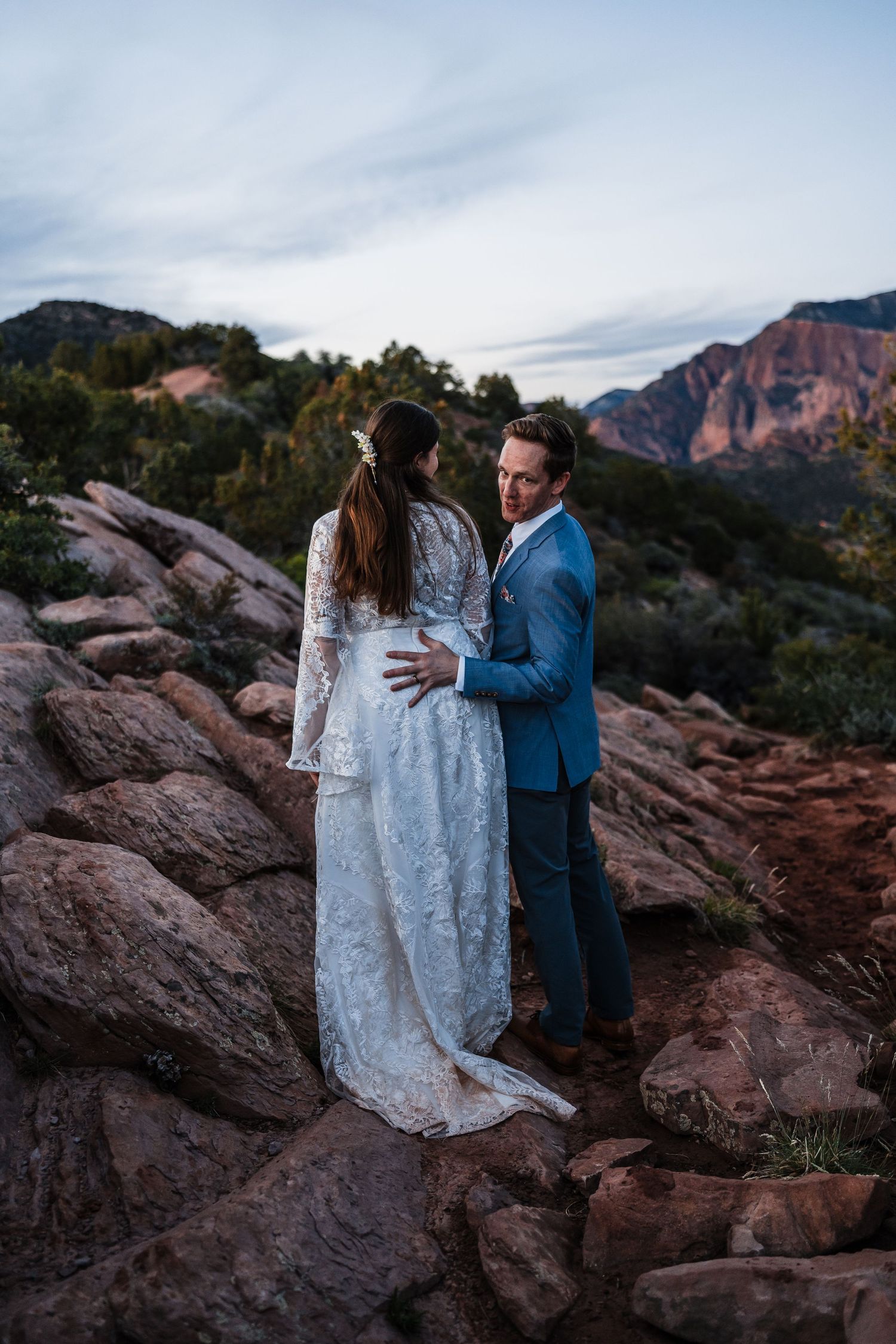Groom in blue suit embracing bride in lace dress on rocky terrain with red mountain backdrop at dusk.