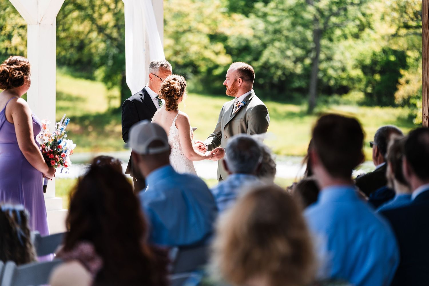 Bride and groom exchange vows at a white gazebo ceremony surrounded by lush green trees and seated guests.
