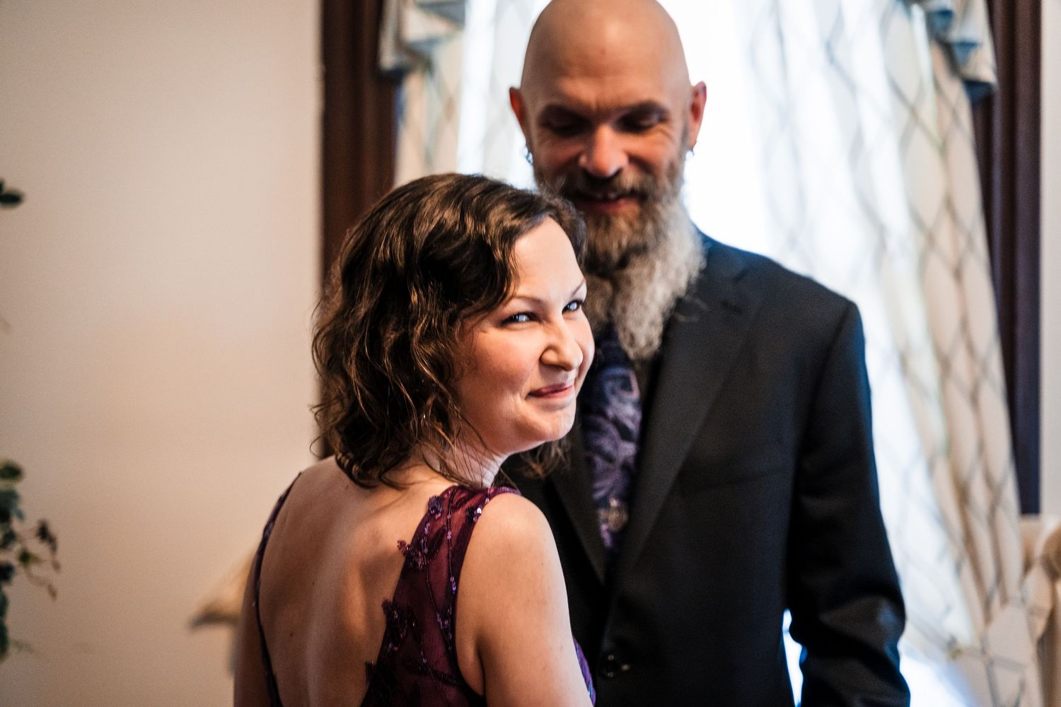 Elegant couple posing indoors, woman in burgundy dress smiling, bald bearded man in dark suit behind her.
