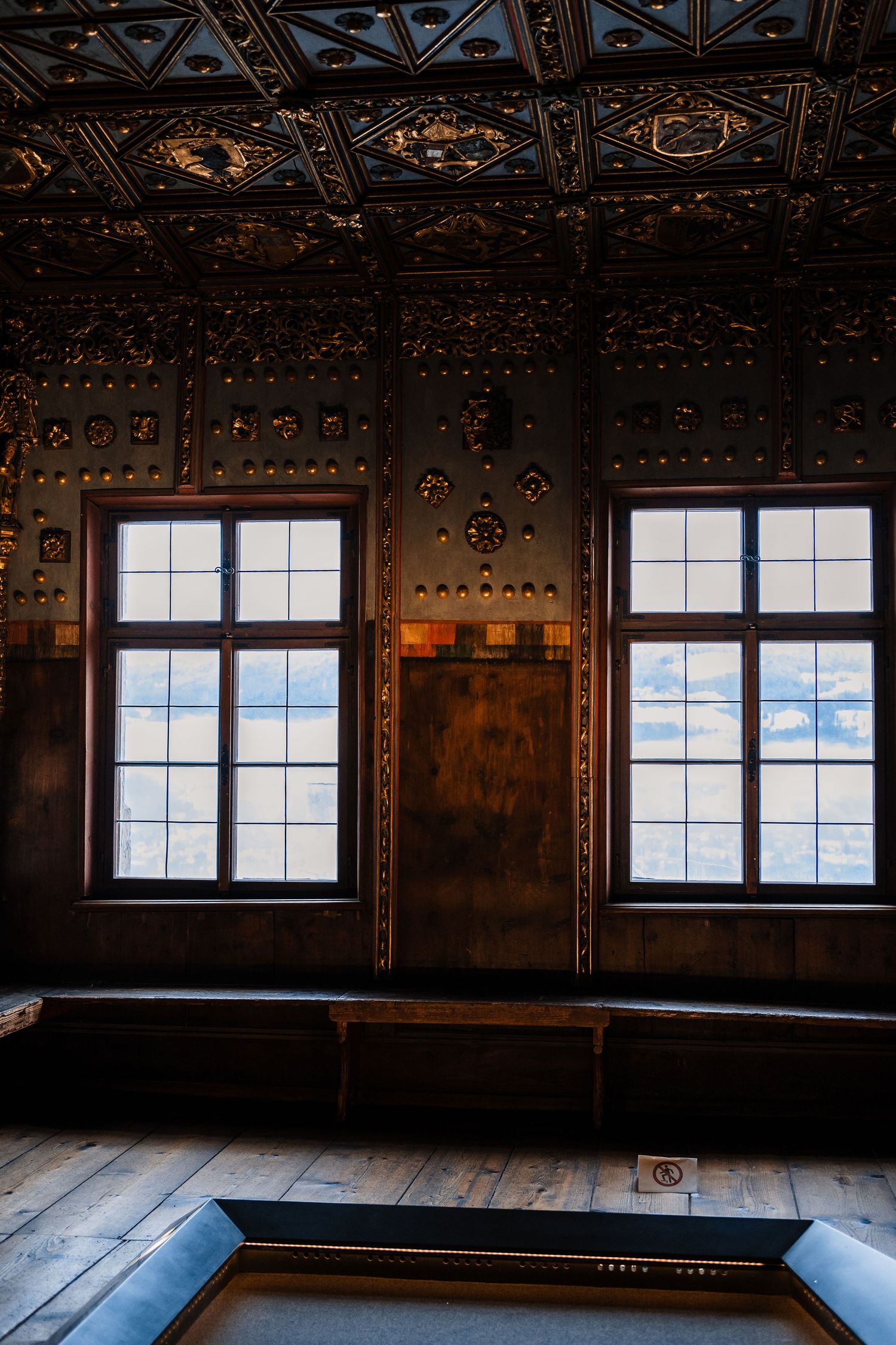 Interior view of a dark wooden room with ornate ceiling details and bright windows letting in natural light.