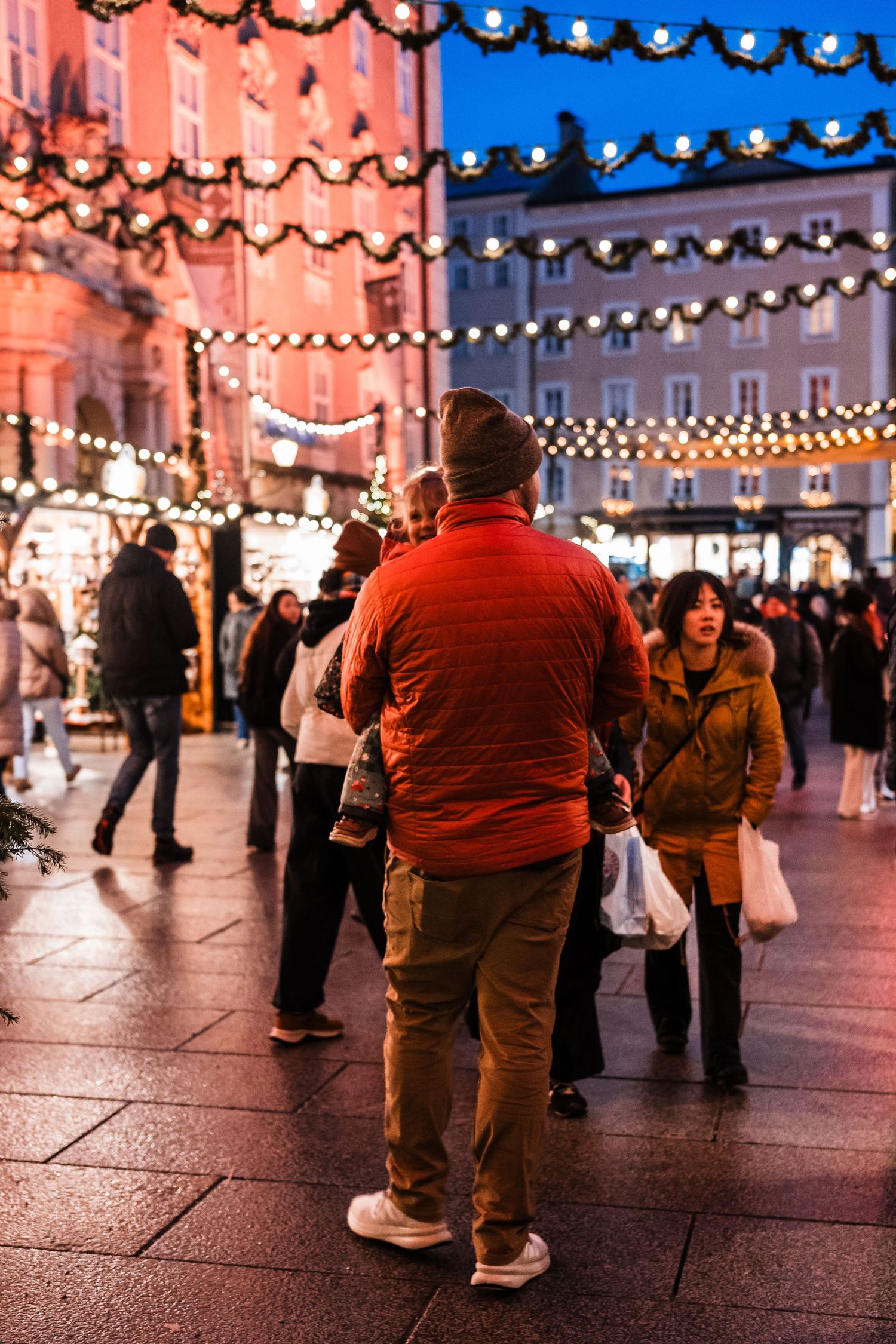People walking through a festive plaza illuminated by overhead string lights at dusk.