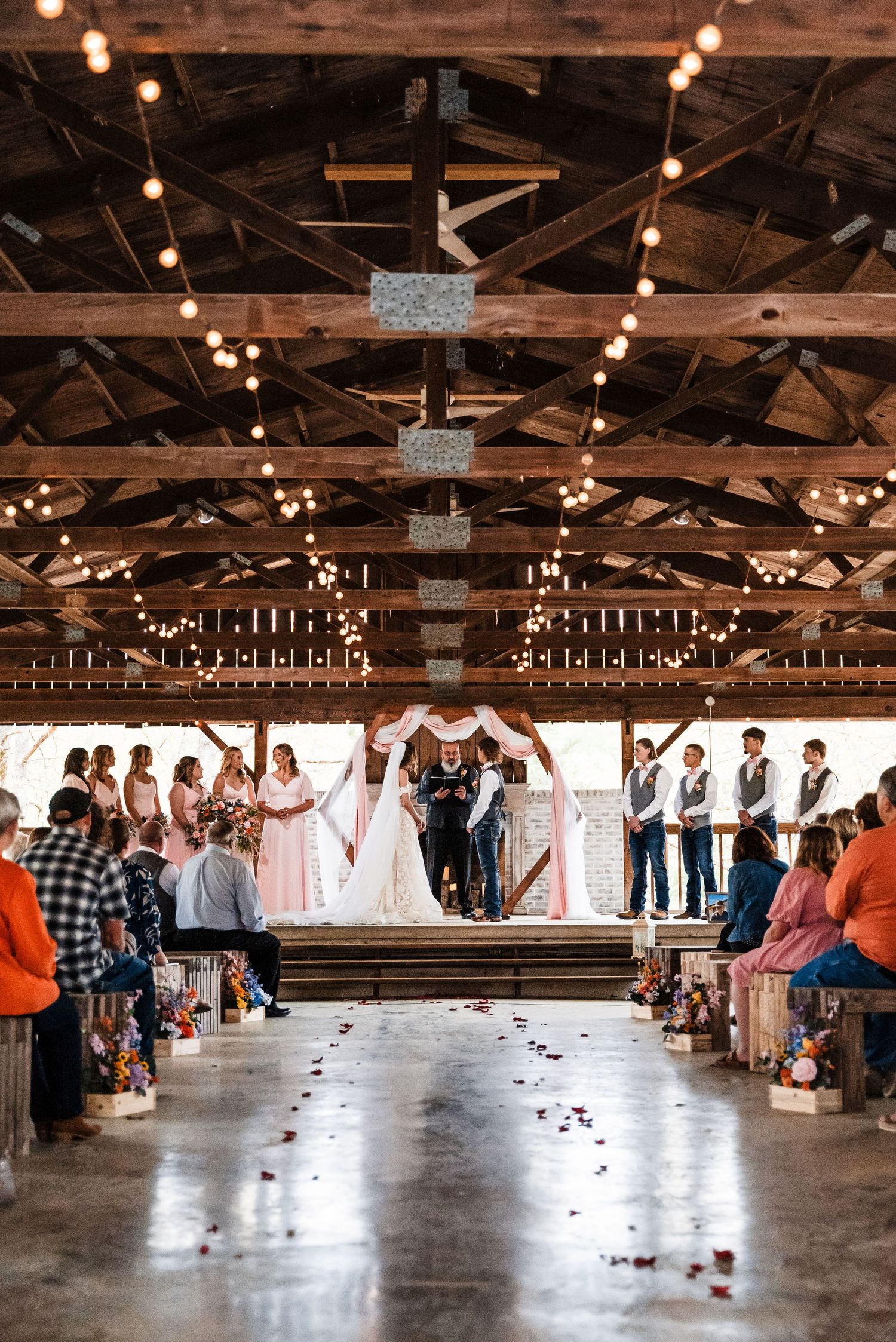 A wide view of a barn wedding ceremony with string lights and wooden beam architecture.
