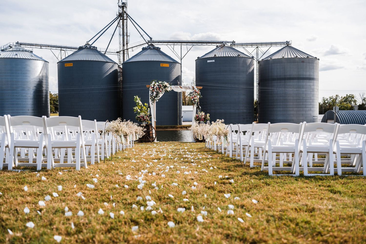 White chairs set up for outdoor wedding ceremony with grain silos in background.