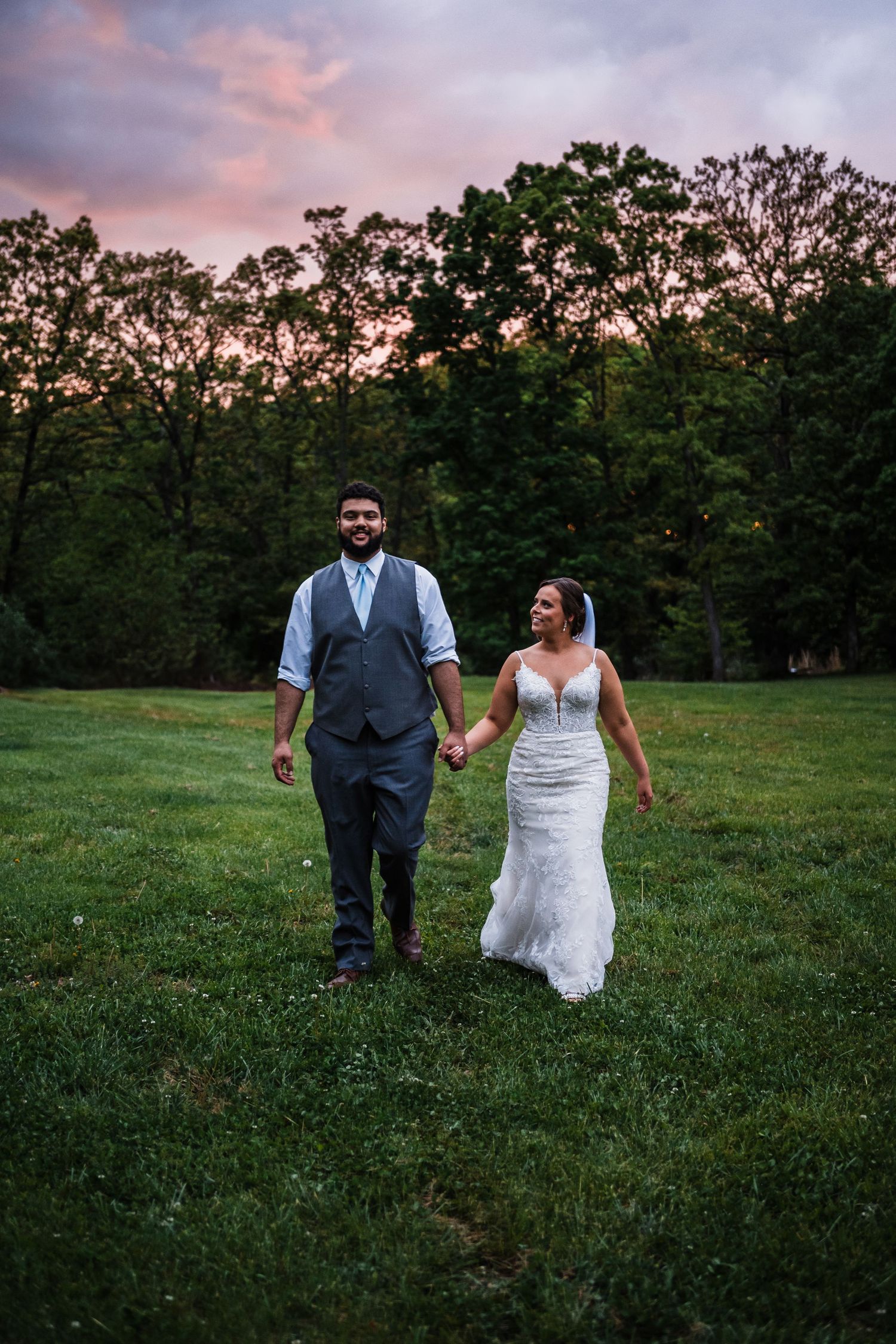A couple walks hand in hand across a grassy field at sunset with trees silhouetted in the background.