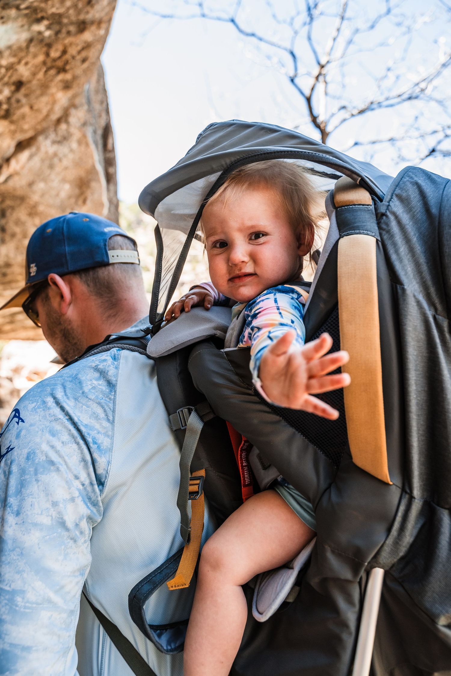 A baby smiles while being carried in a hiking backpack carrier on a sunny day.