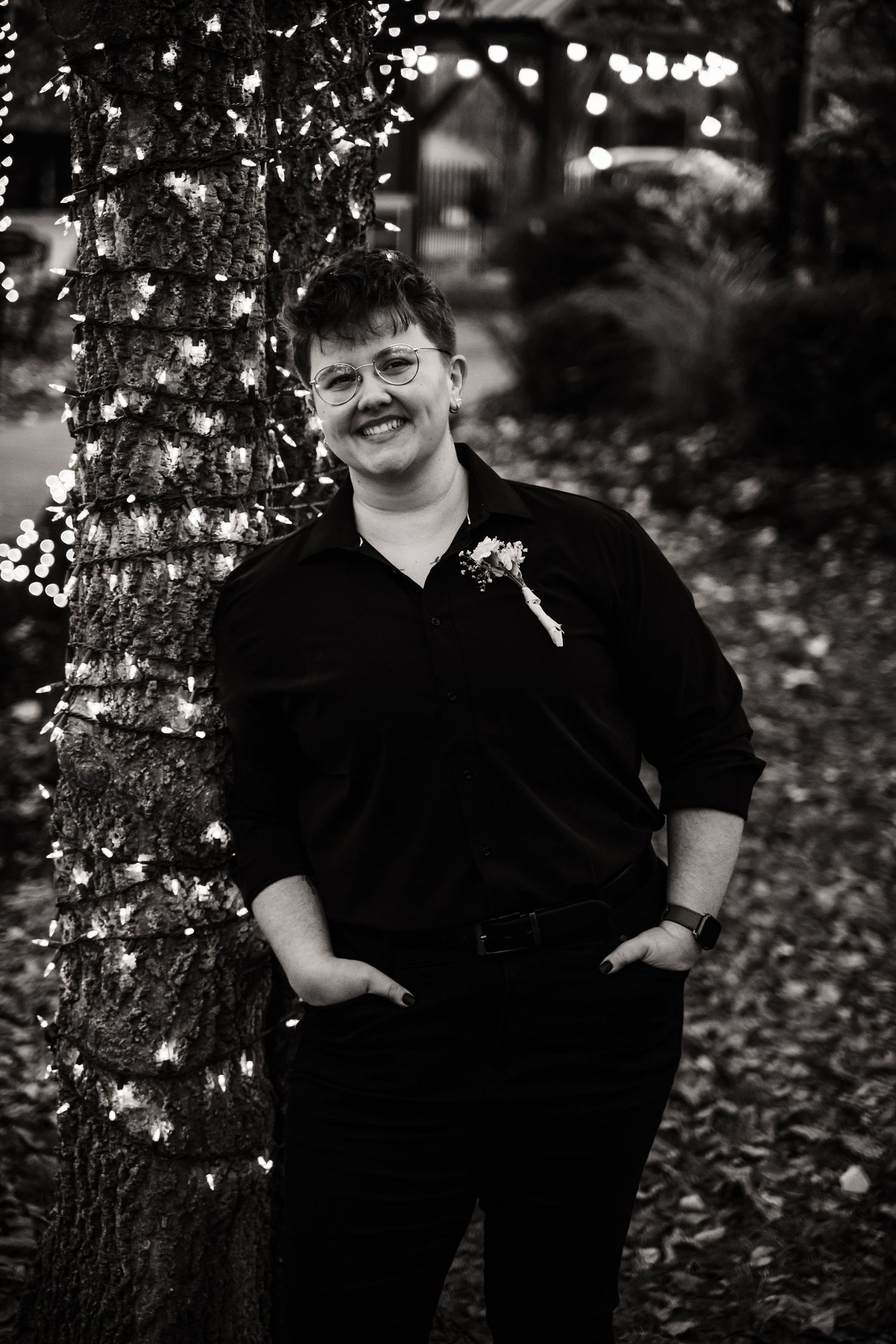 A person in black formal attire with a boutonniere smiles while standing by twinkling lights.