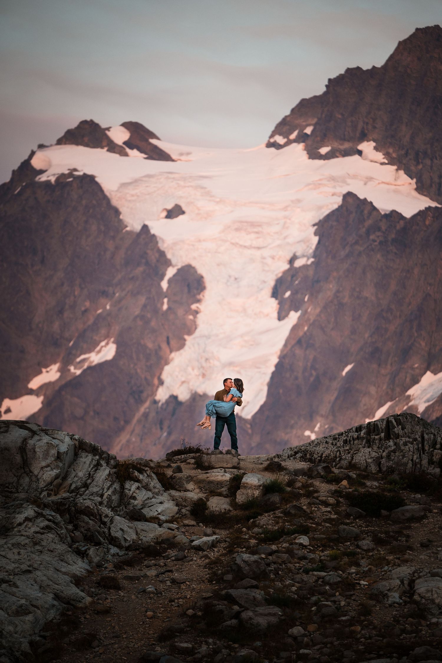 A hiker stands silhouetted against snowy mountain peaks at sunset, creating a dramatic alpine landscape view.