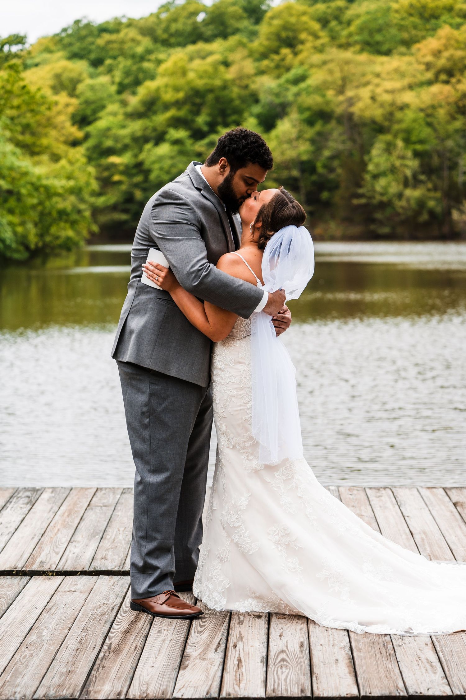 A romantic wedding couple shares an intimate moment on a wooden dock overlooking a serene lake surrounded by lush trees.