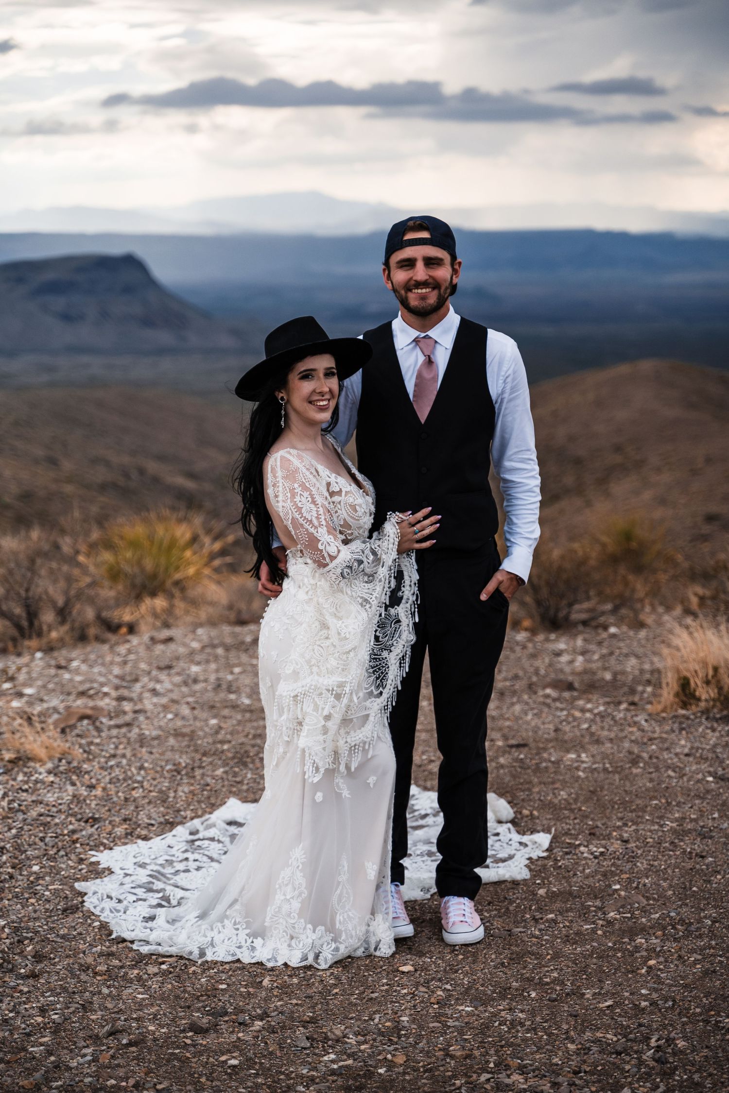 Wedding couple in casual attire poses against dramatic desert mountain landscape.