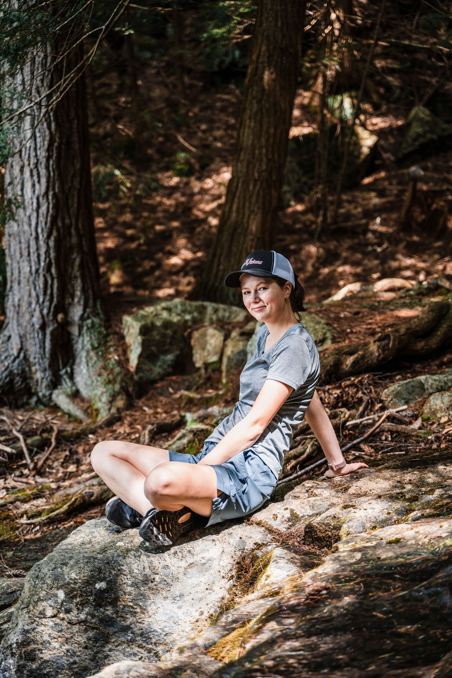 A hiker wearing casual clothing and a baseball cap sits on mossy rocks in a shaded forest setting.